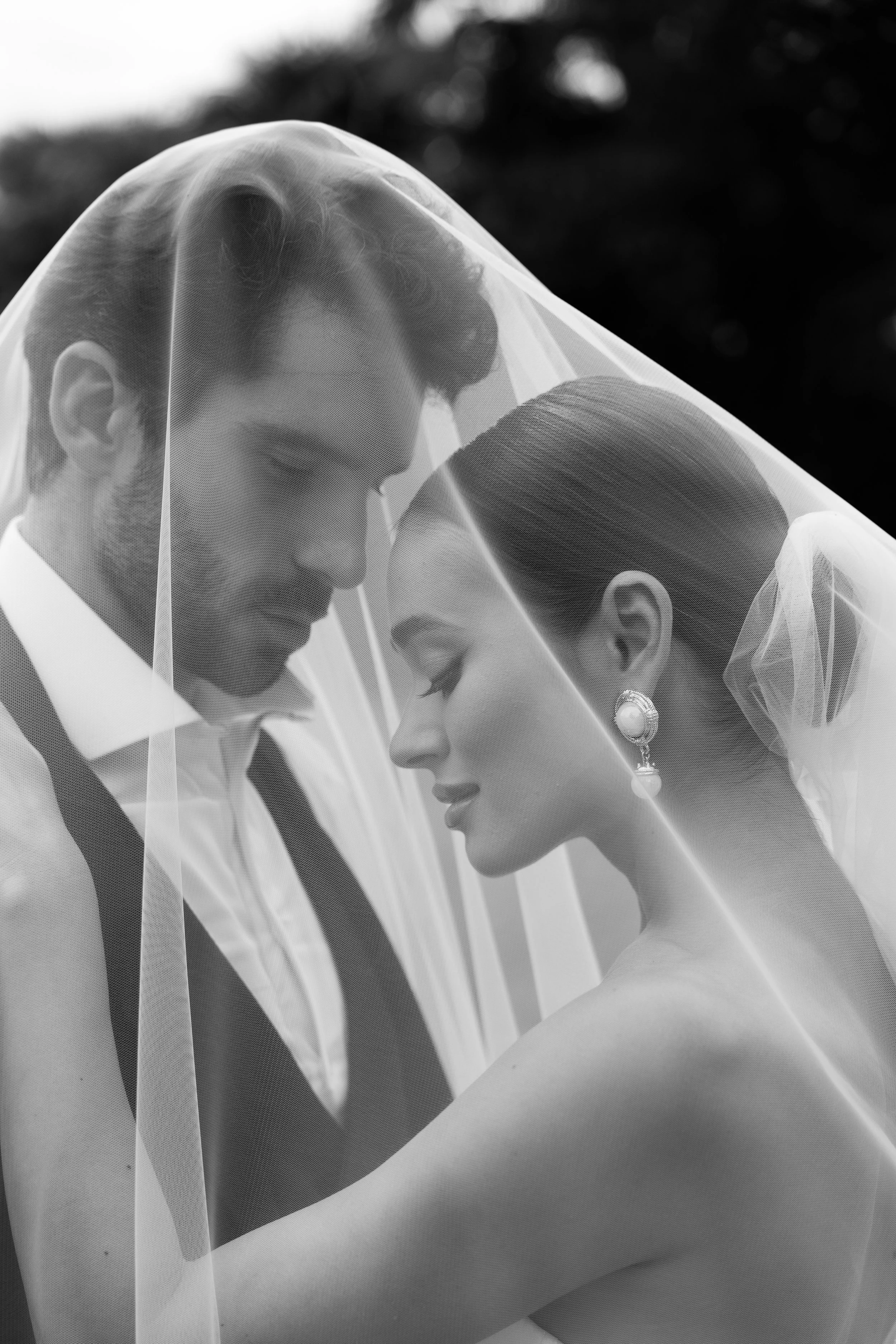 Black and white photograph of a bride and groom with their foreheads touching, smiling softly under a wedding veil.