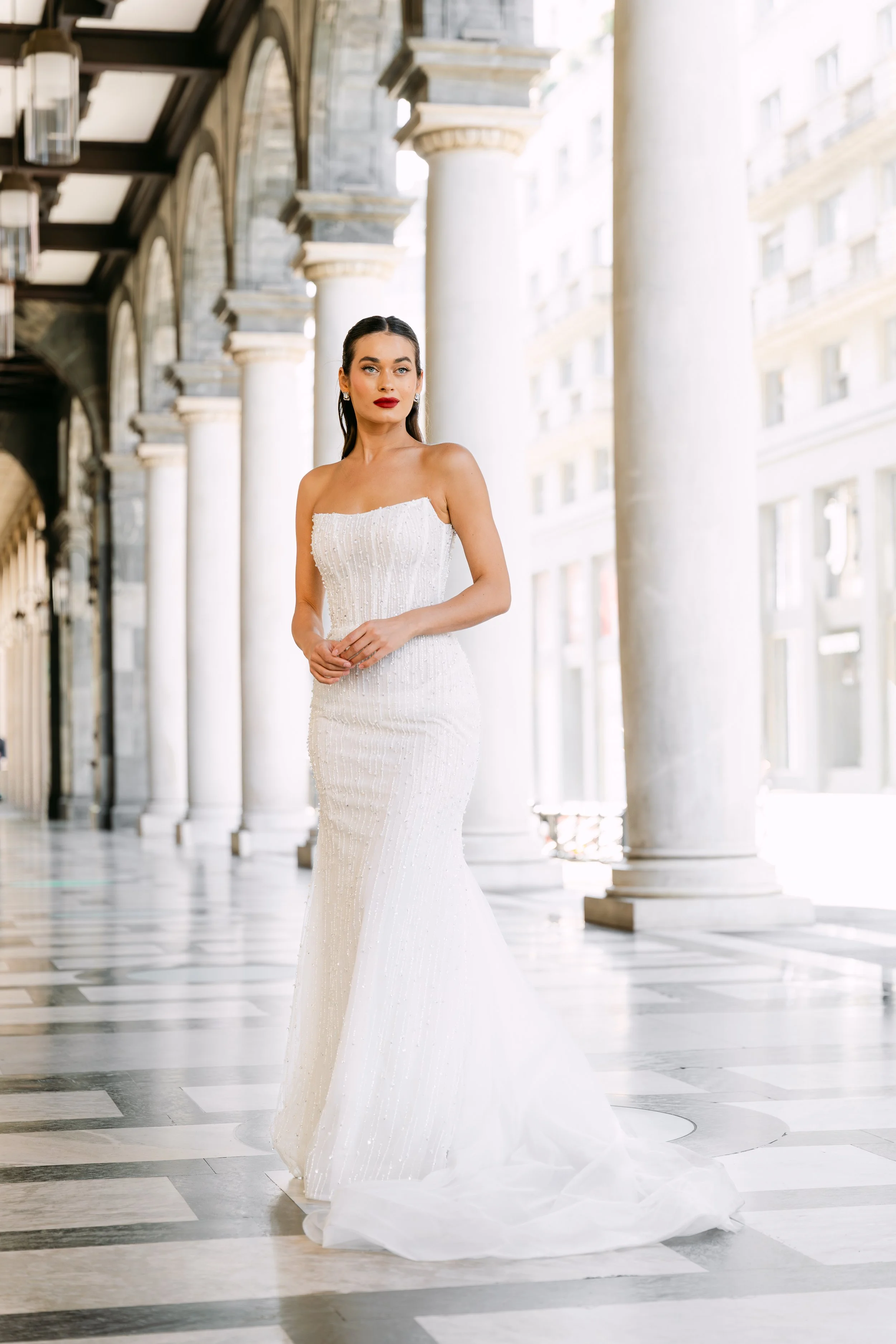 A woman in a white wedding dress with spaghetti straps, standing in an outdoor corridor with tall columns and a black-and-white checkered floor.