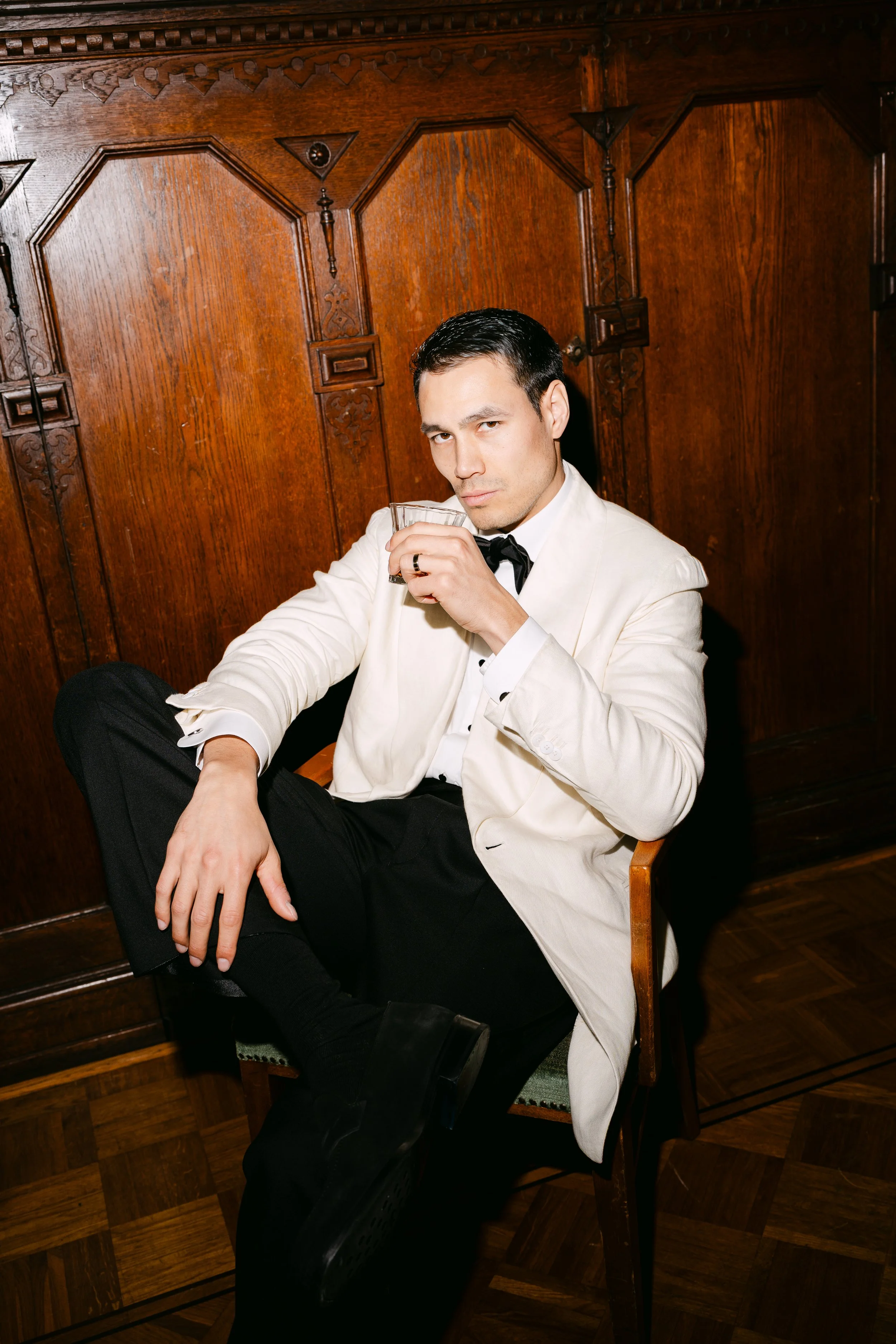 A young man in a white tuxedo jacket and black bow tie, holding a glass, seated on a wooden chair in front of a dark wooden paneled wall.