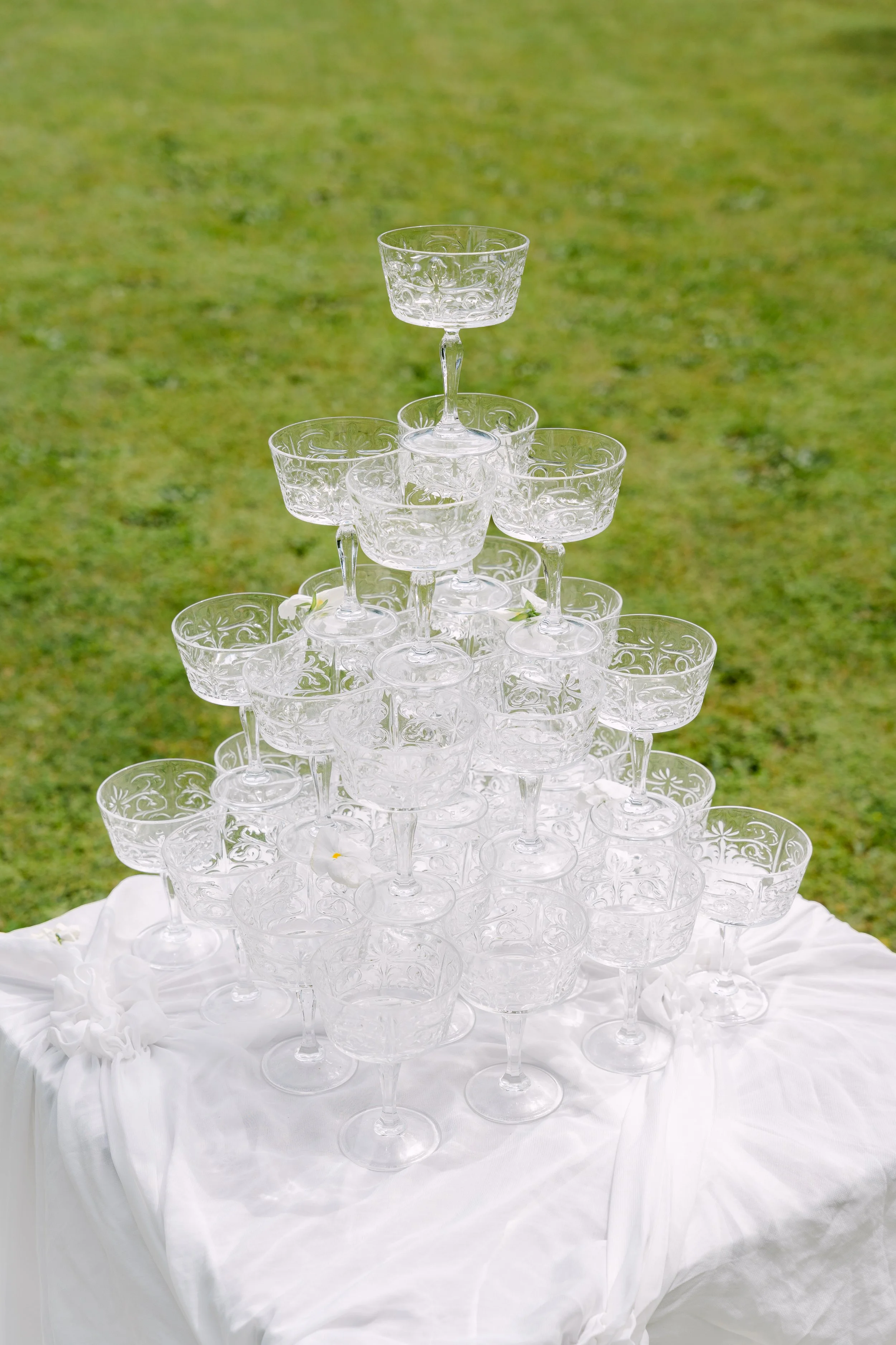 A pyramid of stacked clear glass champagne coupes on a white cloth-covered table outdoors on grass.
