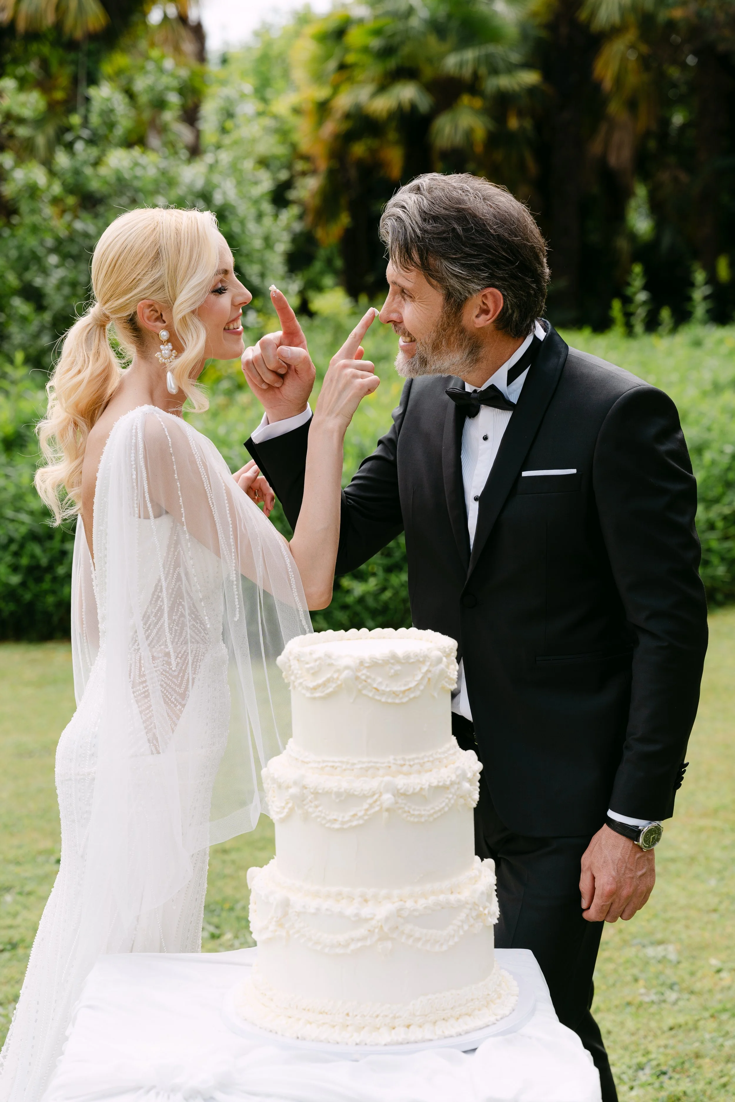 A bride and groom in wedding attire smiling and touching noses during their outdoor wedding, with a large white wedding cake on the table in front of them.