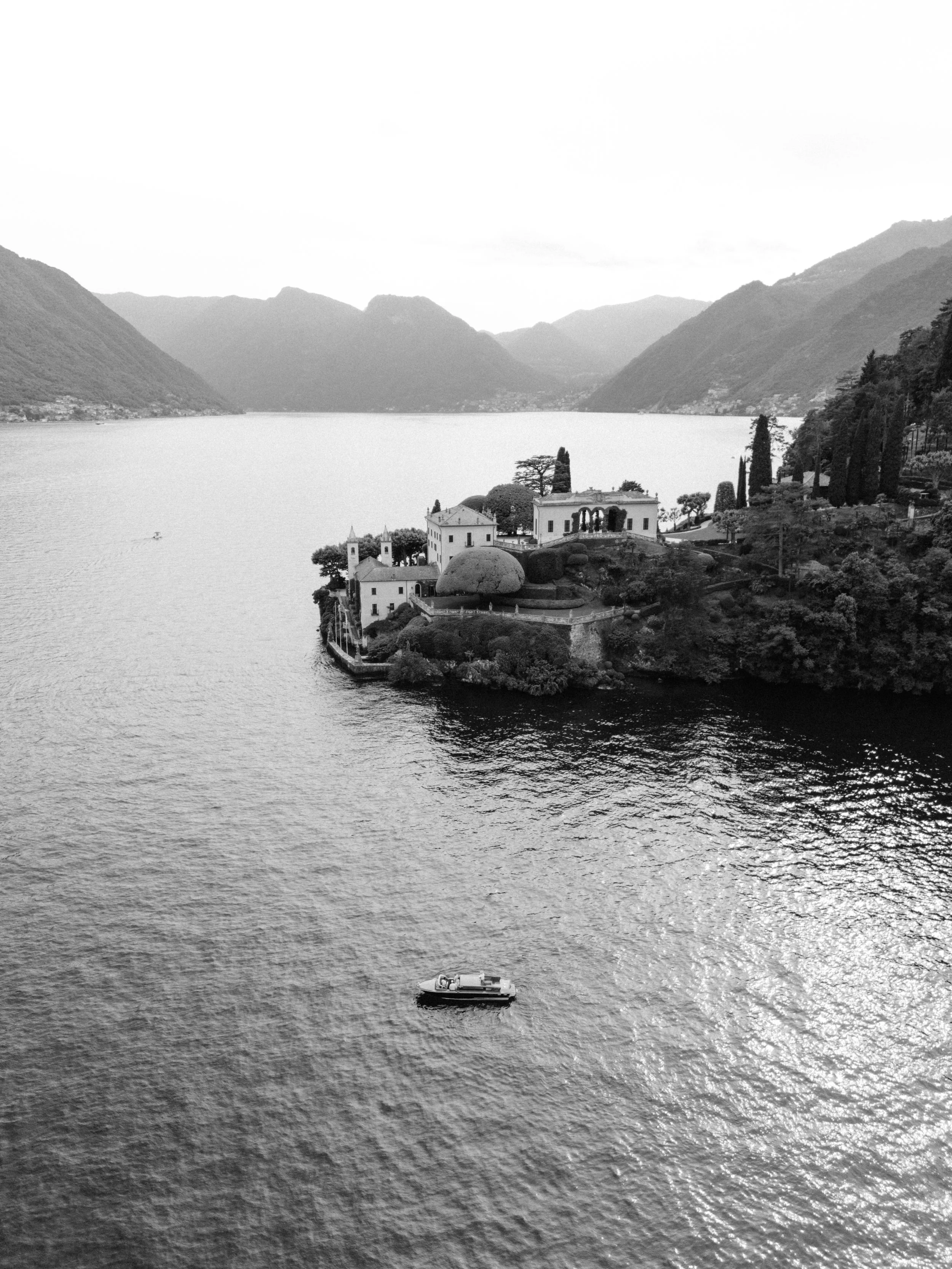 Black and white photo of a lakeside scene showing a small island with a few buildings and tall trees. A boat is floating on the water in the foreground, and hills or mountains are in the background.