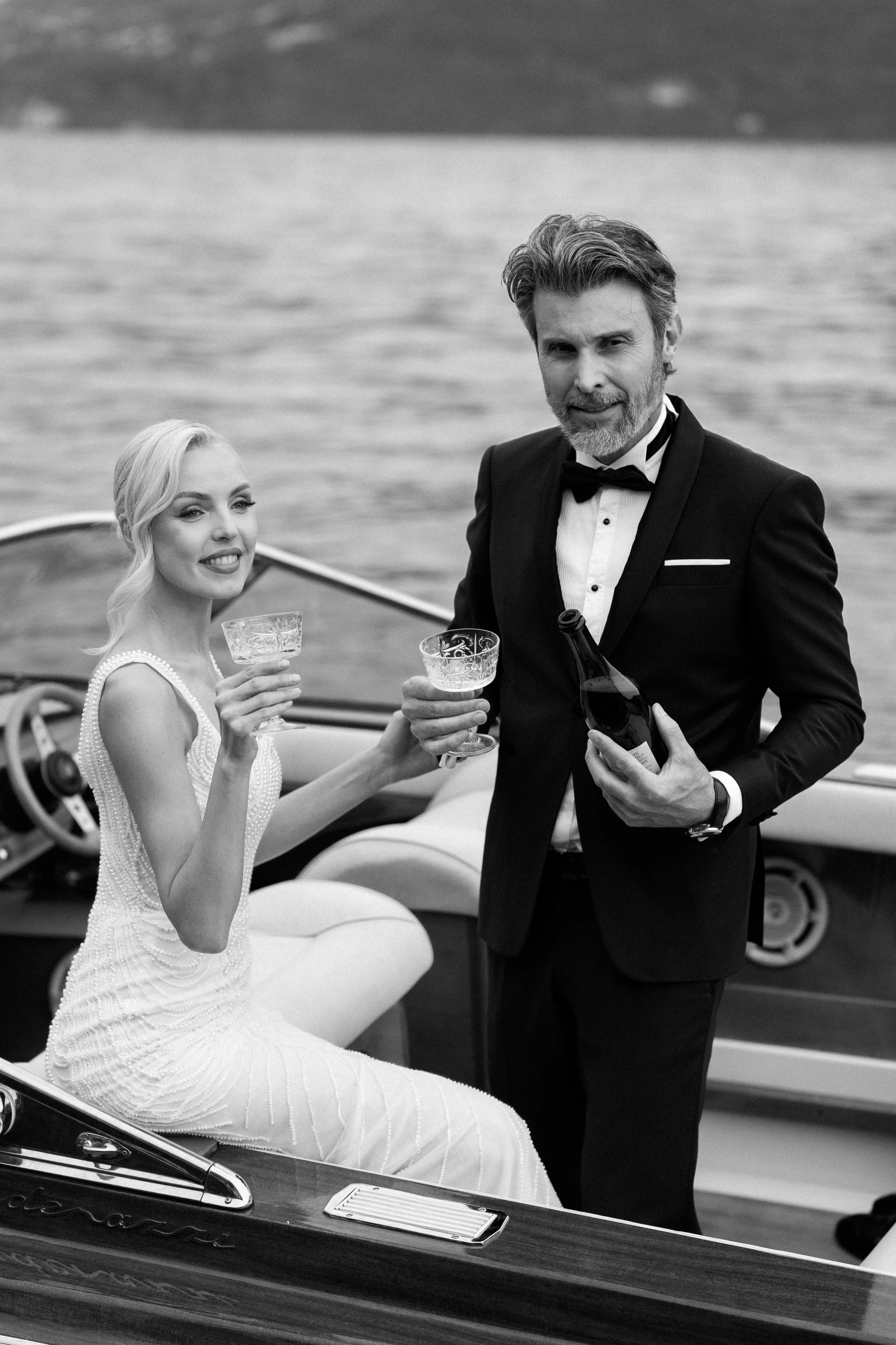 In this black and white photo, a woman in an elegant dress is sitting in a boat, smiling and holding a glass. A man in a tuxedo is standing beside her, holding a drink and a bottle, with an ocean or lake in the background.