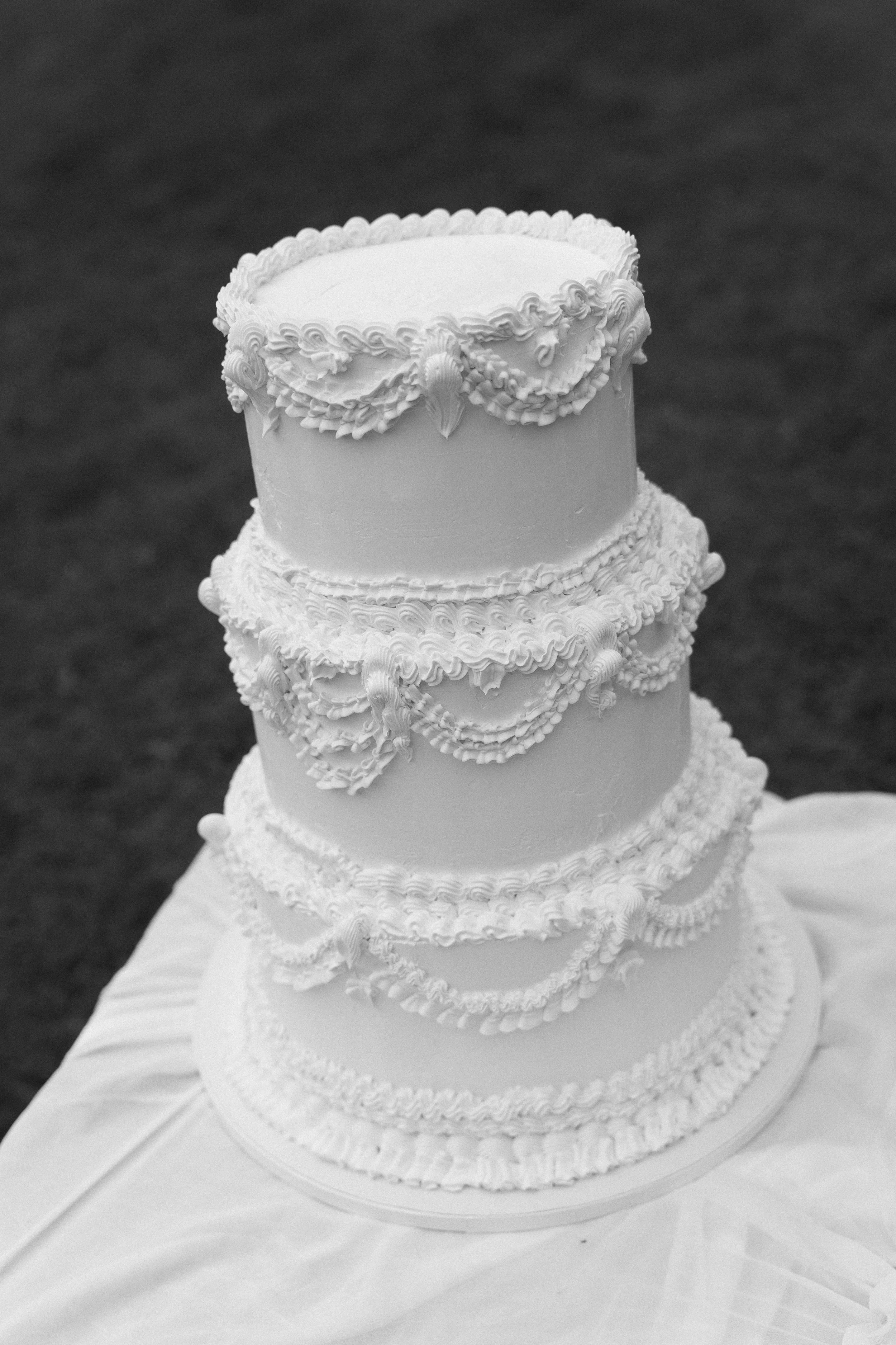 A tall, multi-tiered wedding cake decorated with intricate piping and icing details, placed on a table covered with a cloth.