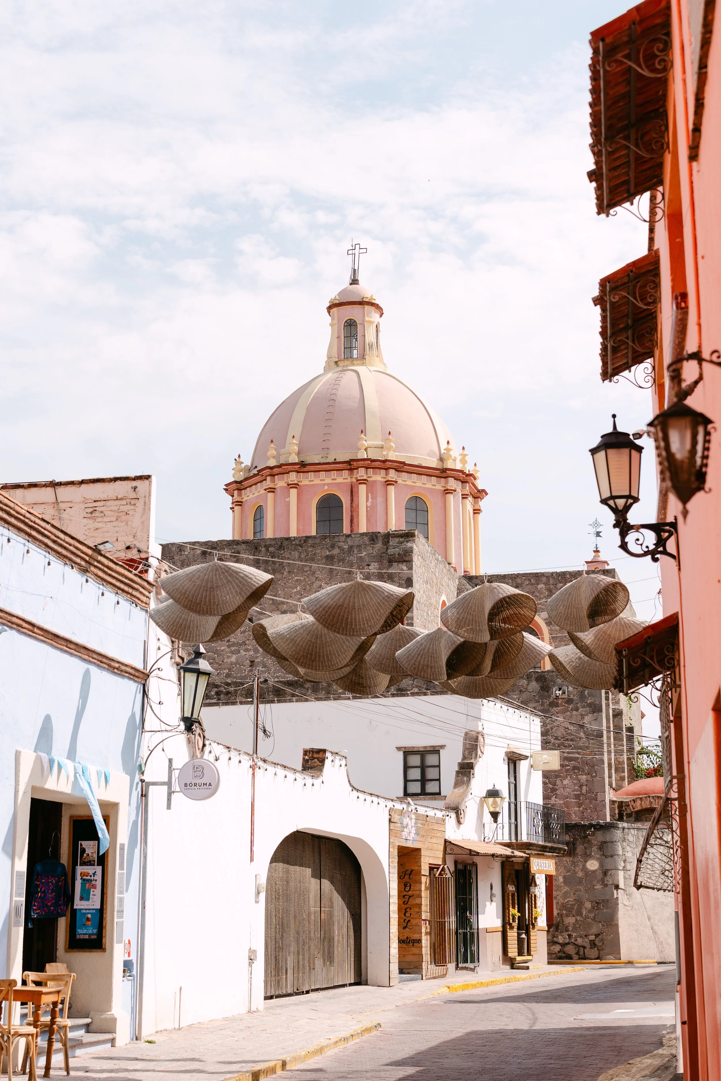 A street scene with colorful buildings, hanging straw hats, and a large pink domed church in the background.
