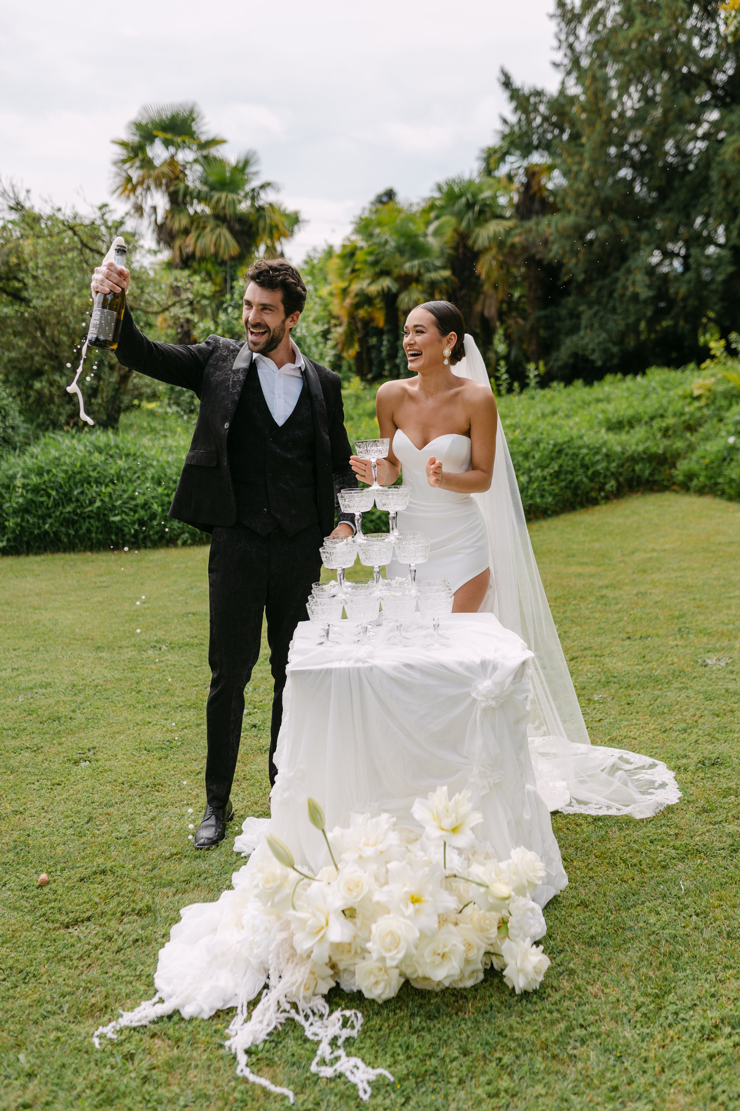 A newly married couple celebrating outdoors with a champagne tower, the groom opening a champagne bottle and the bride smiling, surrounded by flowers and greenery.