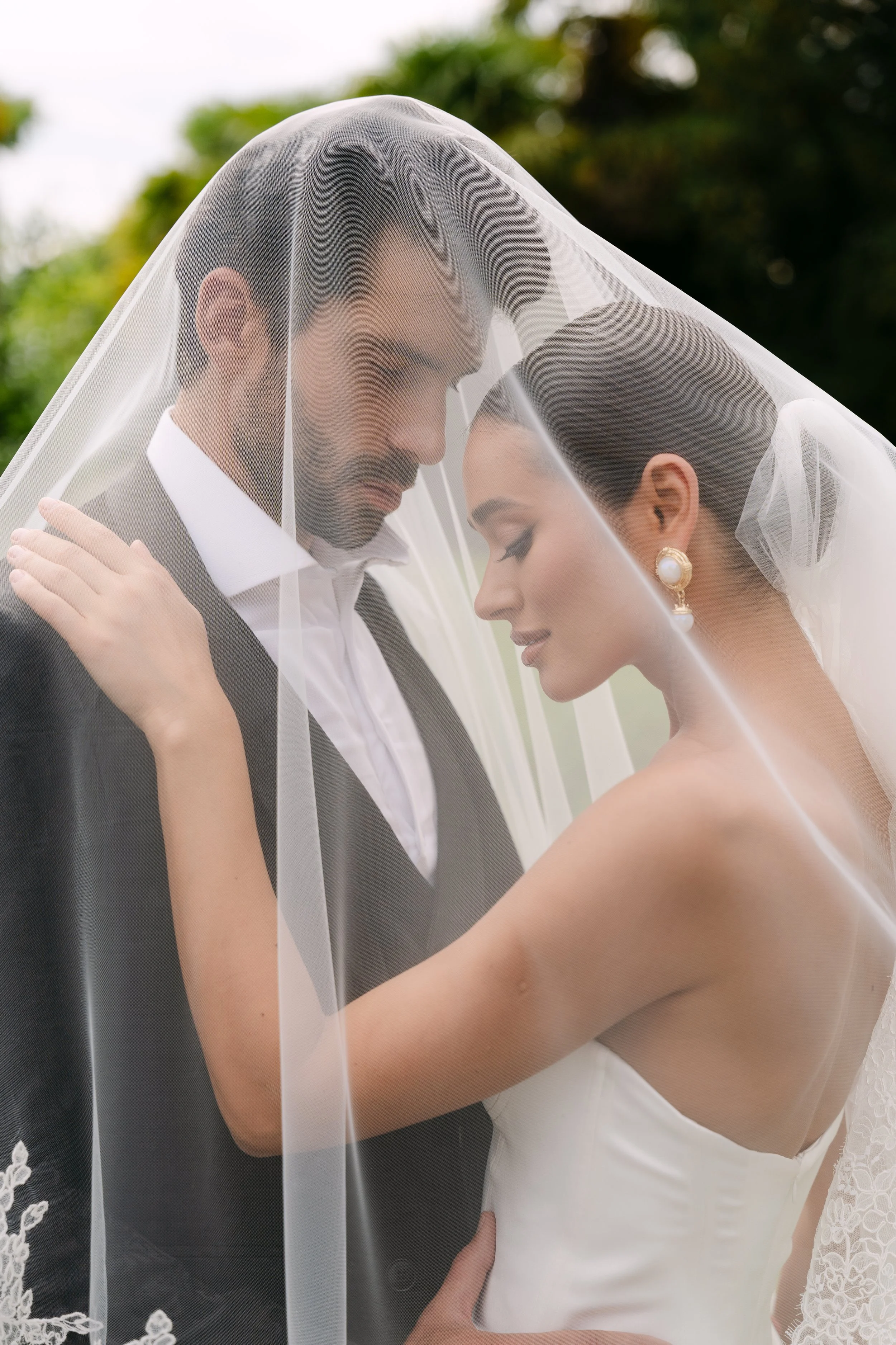 A bride and groom under a wedding veil, gazing at each other, outdoors with greenery in the background.