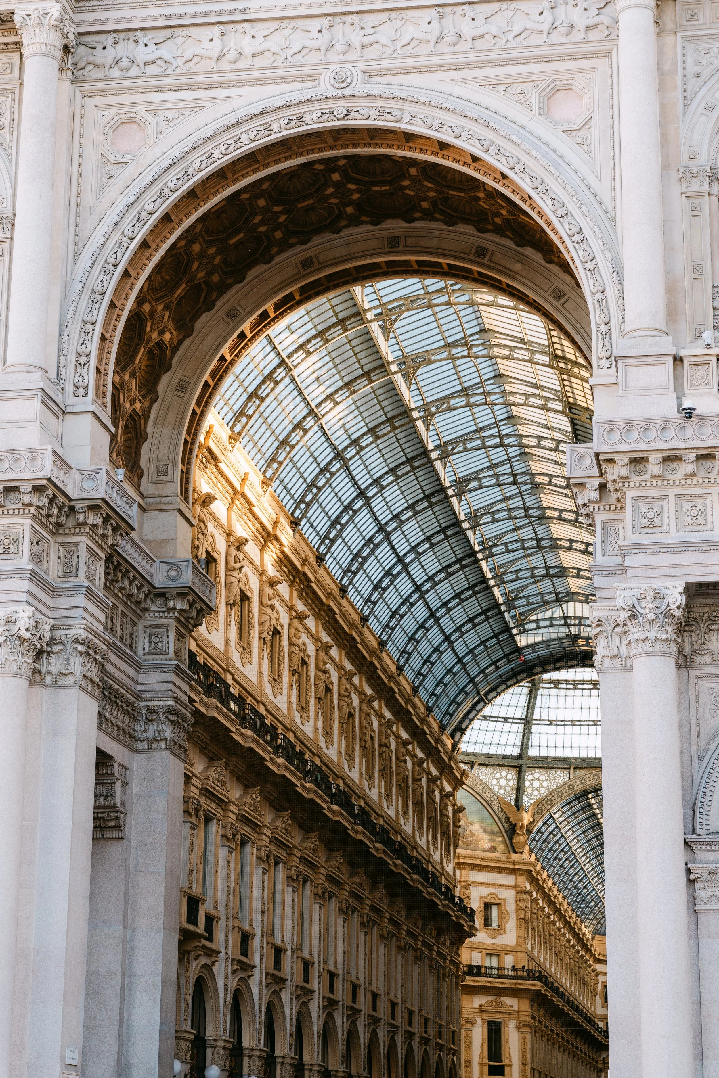 Interior of a grand historic shopping gallery with ornate stone archways, decorative columns, and a curved glass roof allowing natural light.