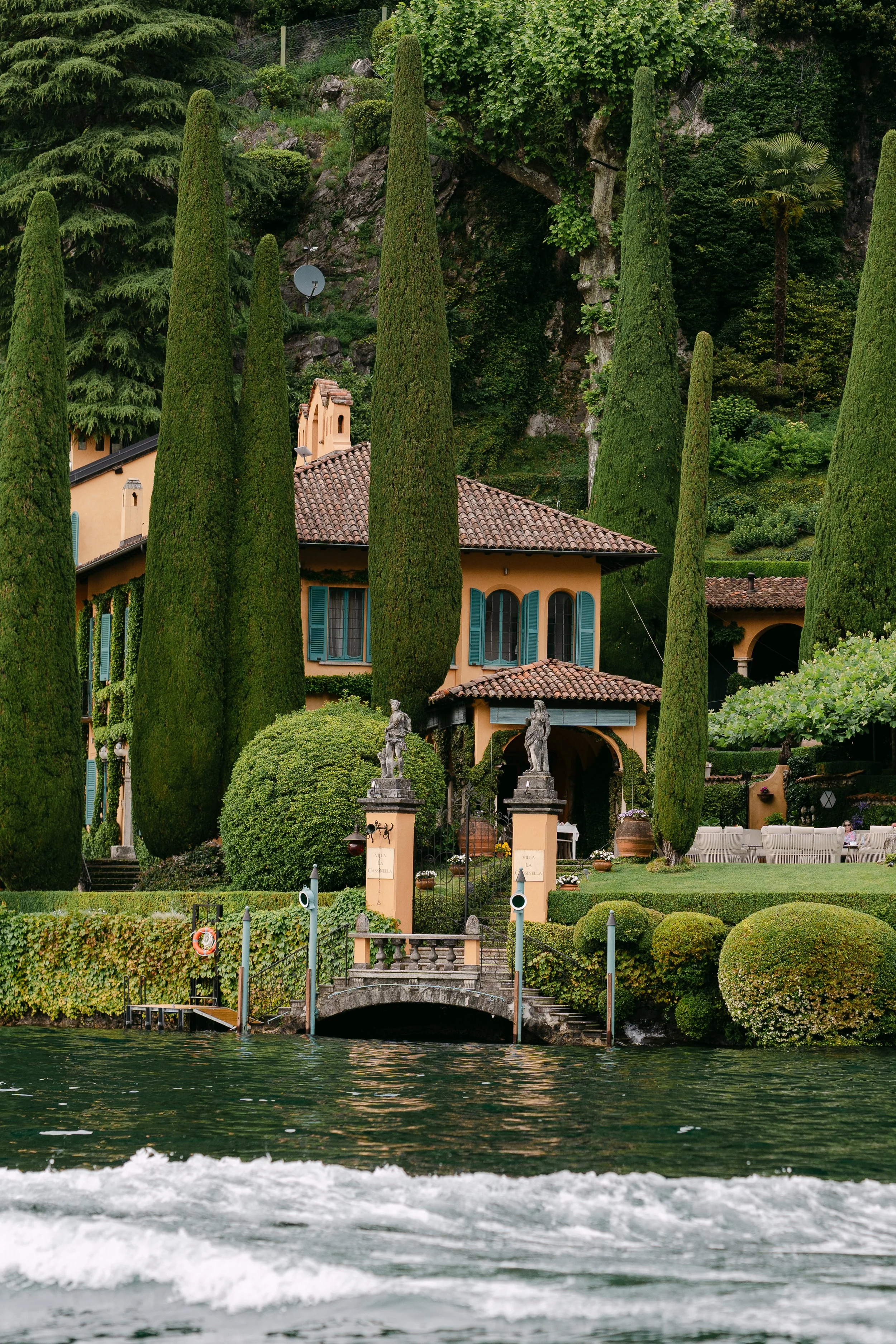 A luxurious lakeside villa surrounded by tall cypress trees and lush greenery, viewed from the water with a small stone bridge in front.