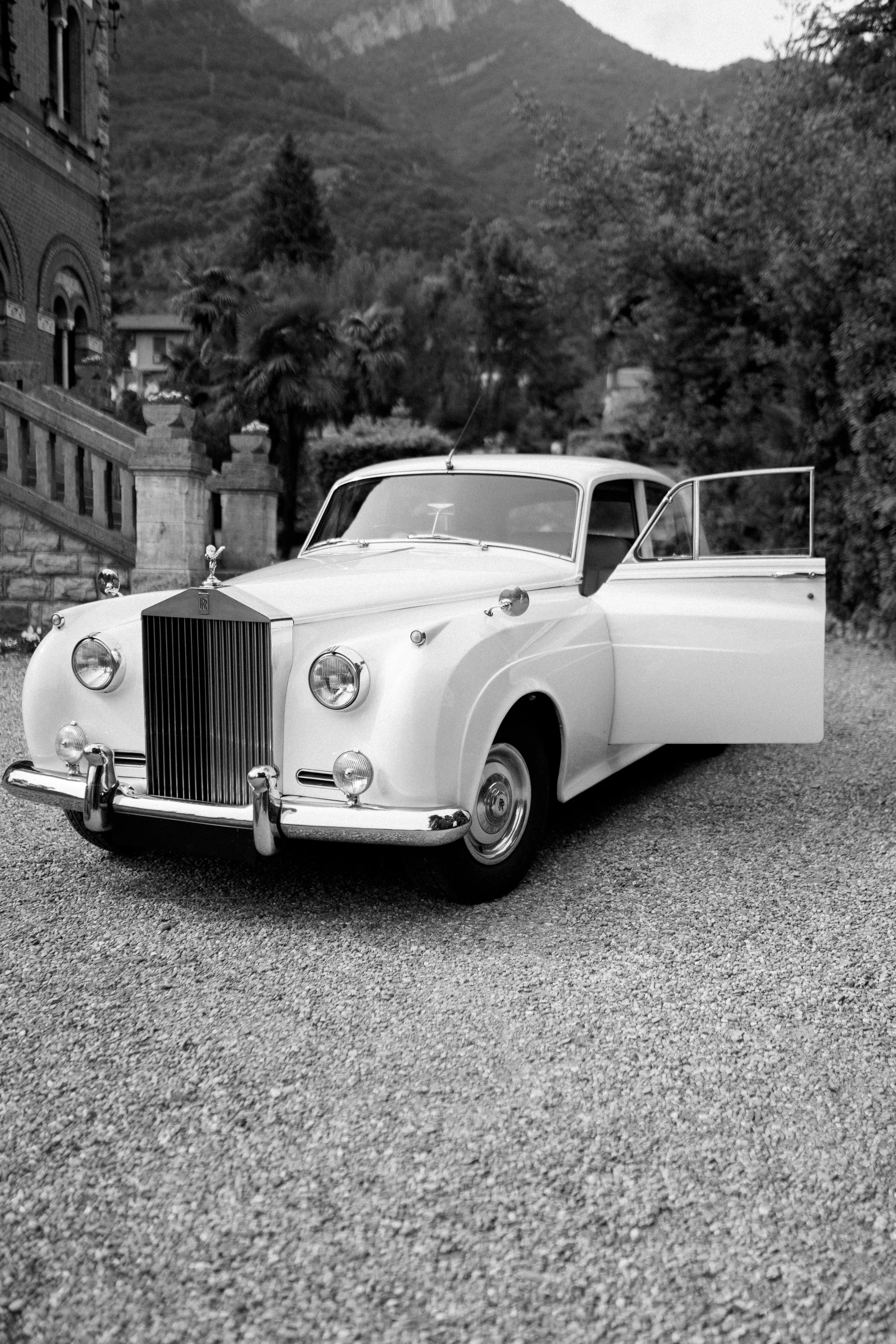 A vintage white luxury car with a distinctive grille, round headlights, and chrome accents parked on a gravel driveway with a historic building, trees, and mountains in the background.