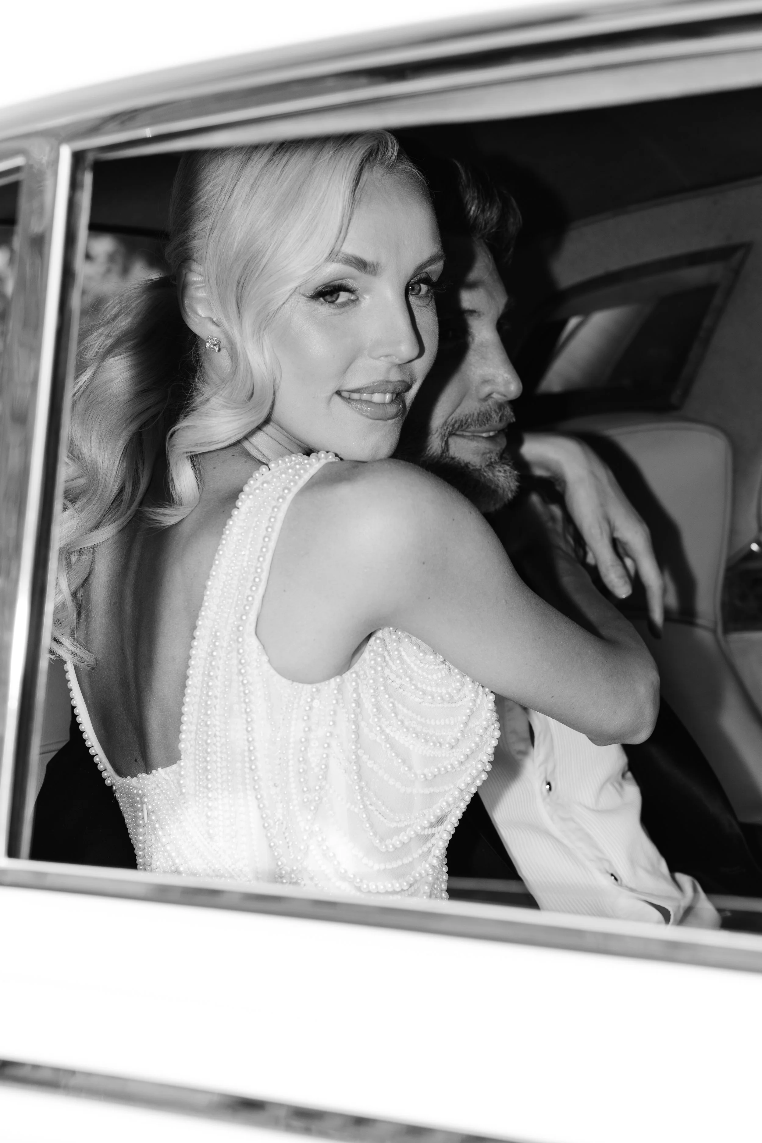 A black-and-white photo of a woman with platinum blonde hair and a man with dark hair and a beard, sitting inside a vintage car, embracing each other.