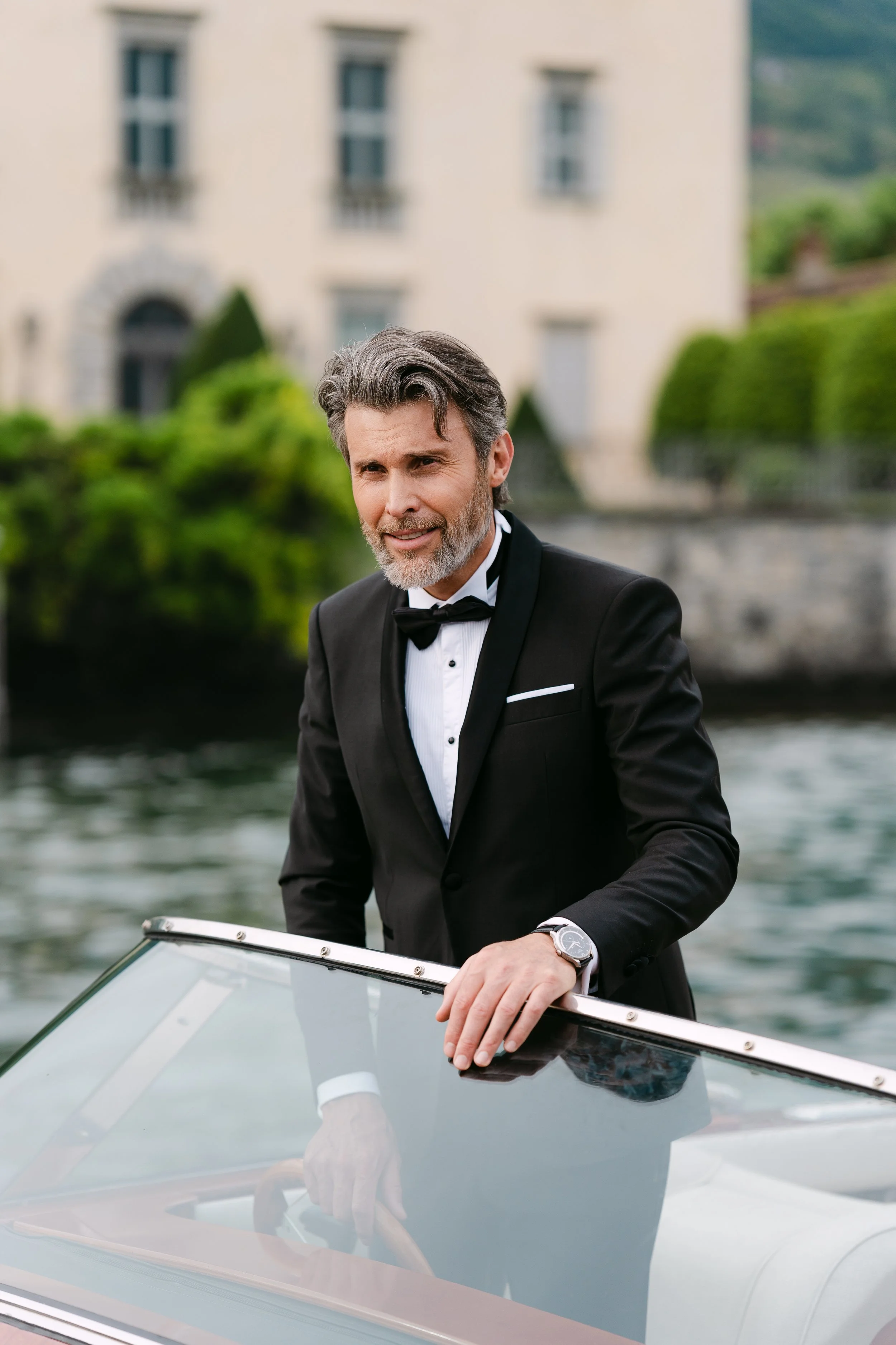 A middle-aged man with gray hair and beard in a tuxedo standing on a boat, holding the helm, during daytime.