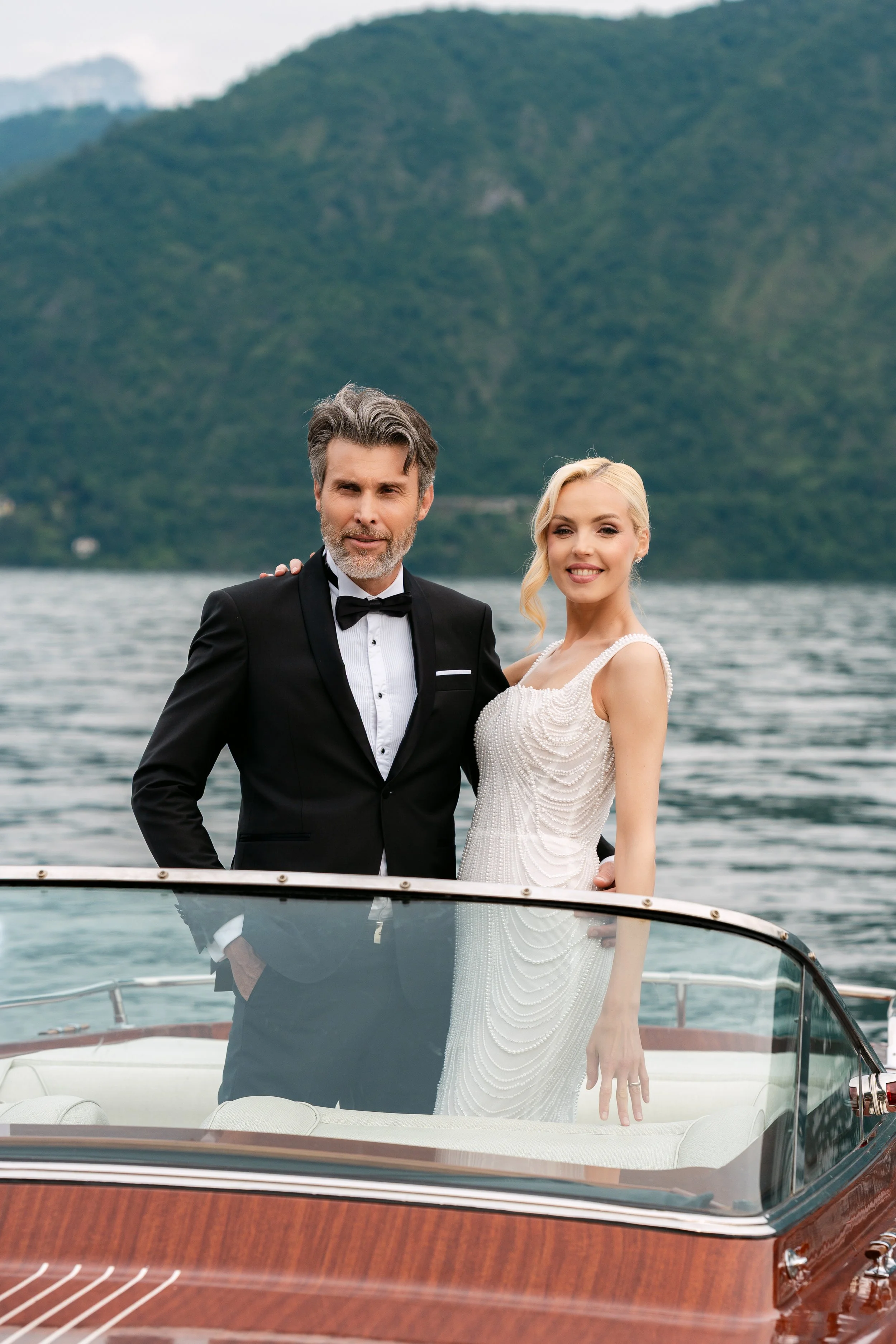 A bride and groom standing on a boat with water and green mountains in the background, dressed in wedding attire.
