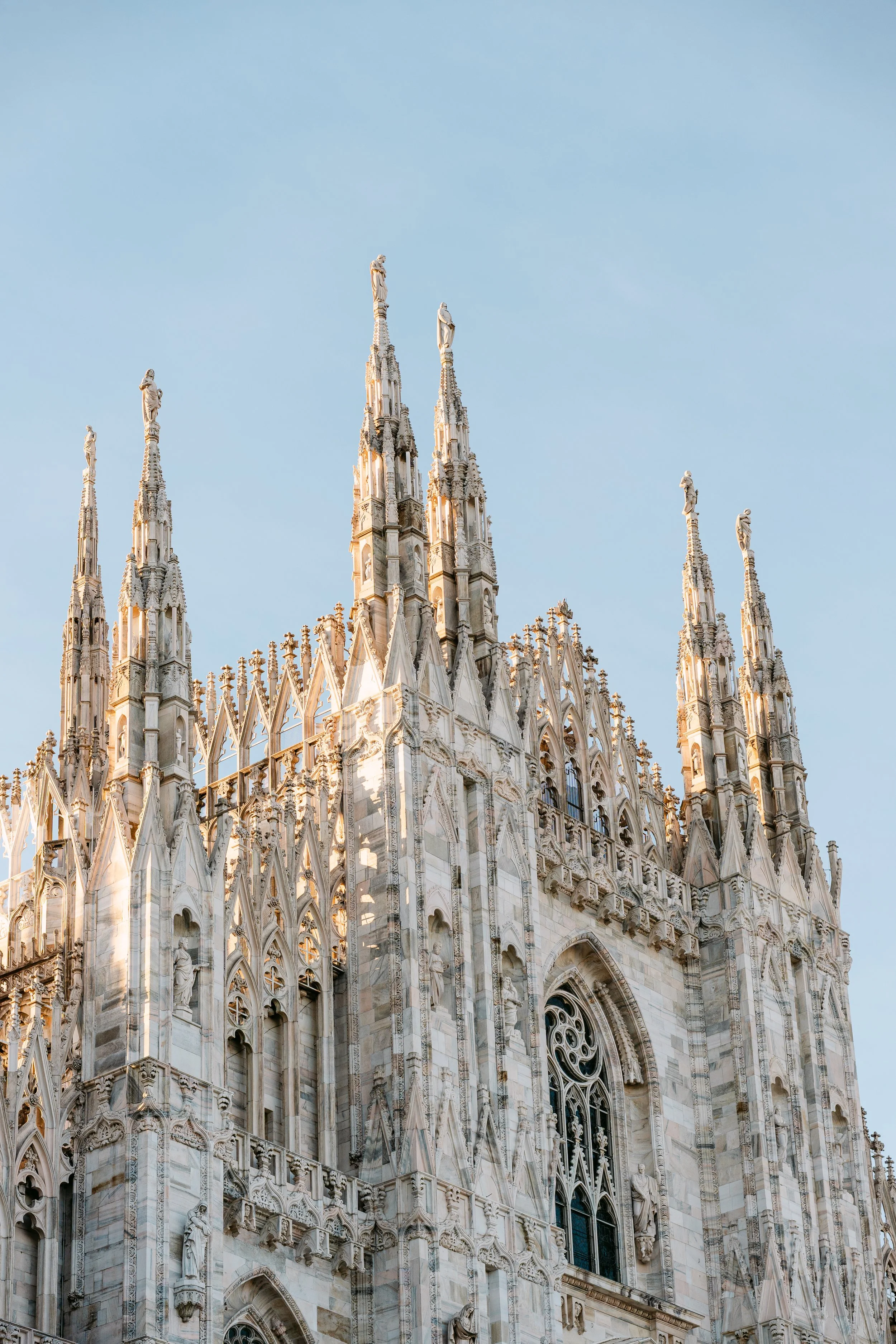 Close-up of the ornate, gothic-style upper facade of a white marble cathedral with intricate carvings and statues against a clear, blue sky.