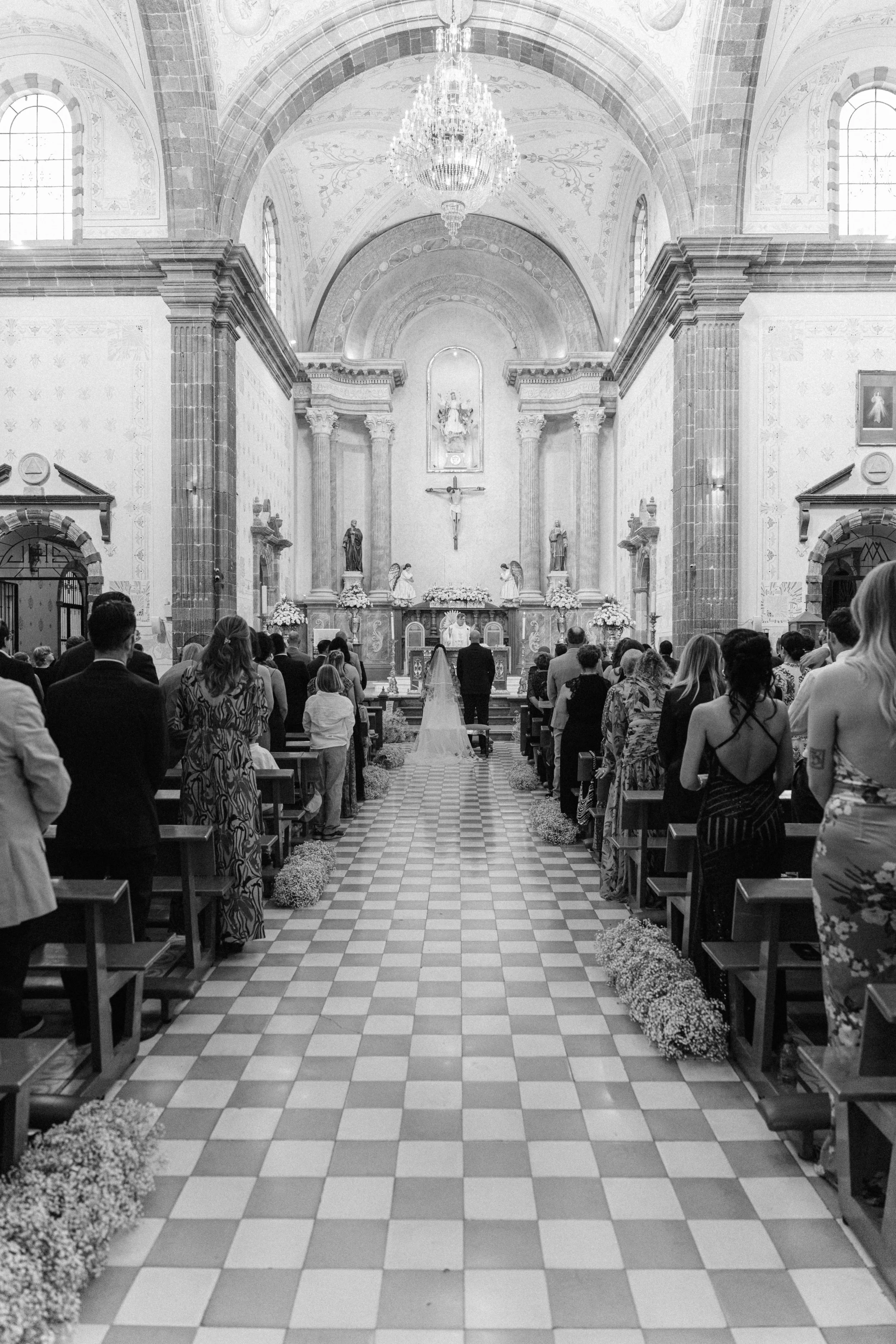 A black and white photo of a wedding ceremony inside a church with a high arched ceiling, chandeliers, and religious statues. The bride and groom are standing at the altar, facing the officiant, while guests are seated on wooden pews on either side o