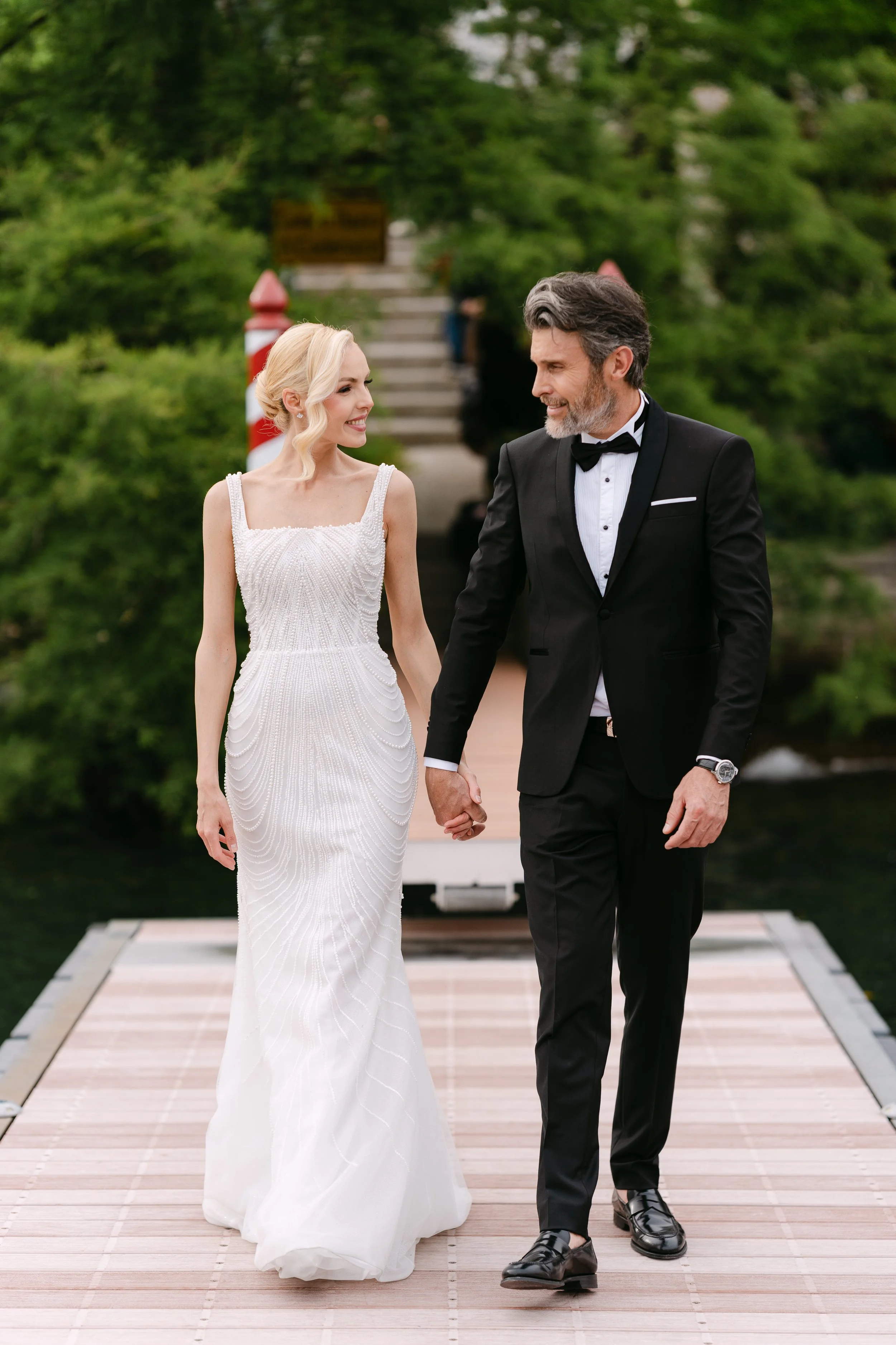 A bride and groom walking hand in hand on a wooden dock, surrounded by green trees, during their wedding ceremony.