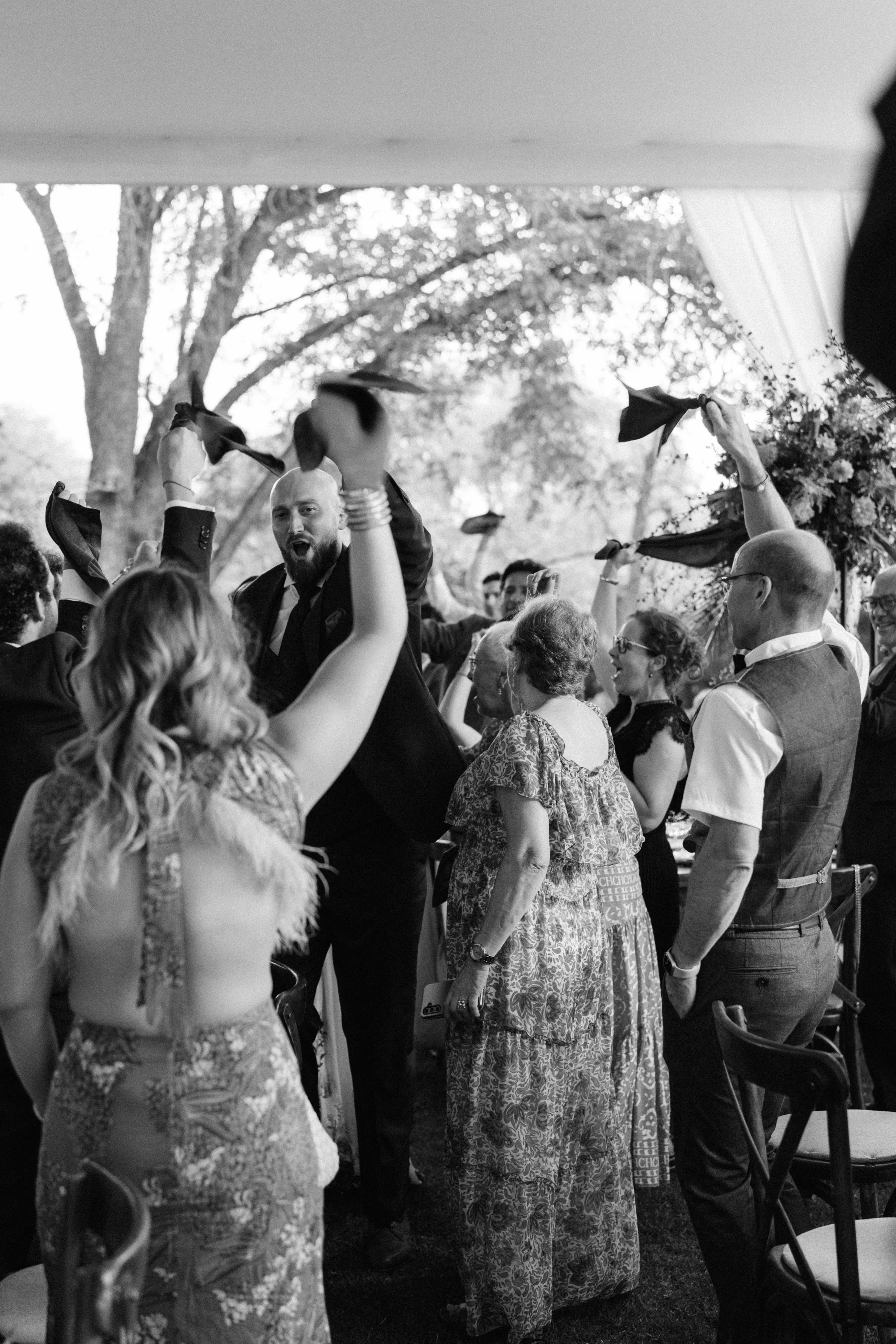 People celebrating at a wedding reception, waving cloths and smiling. It is outdoors, with trees in the background.