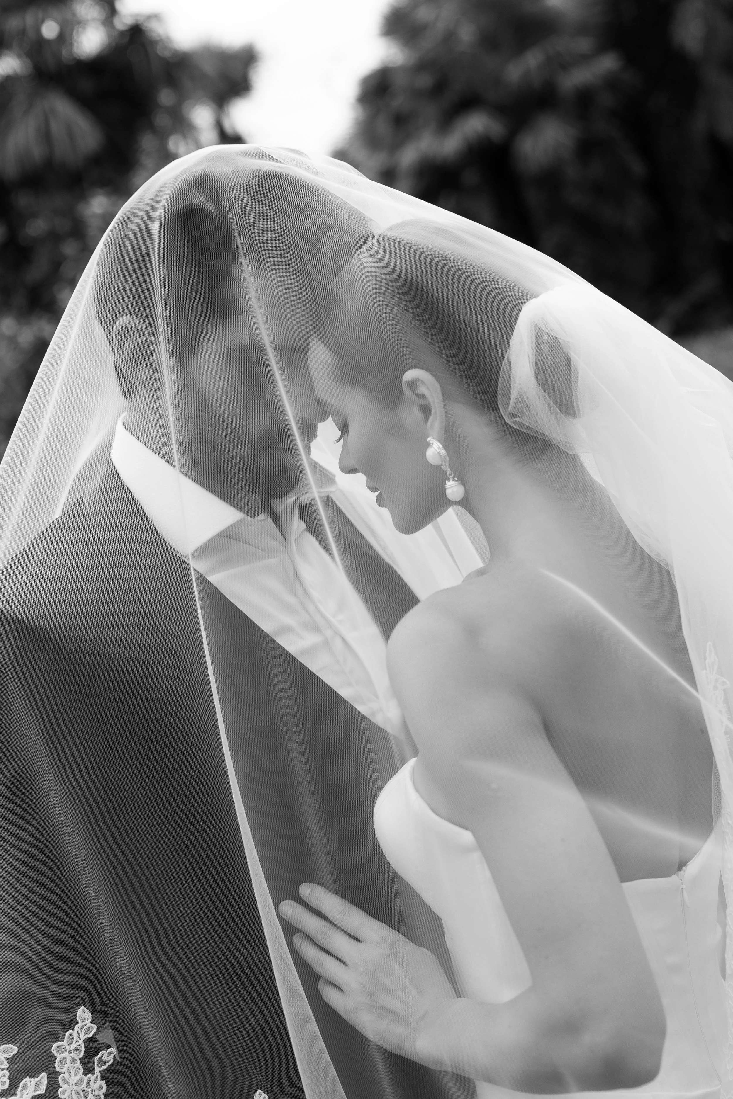 Black and white photo of a bride and groom sharing an intimate moment under a veil, with their foreheads touching, outdoors with blurred trees in the background.