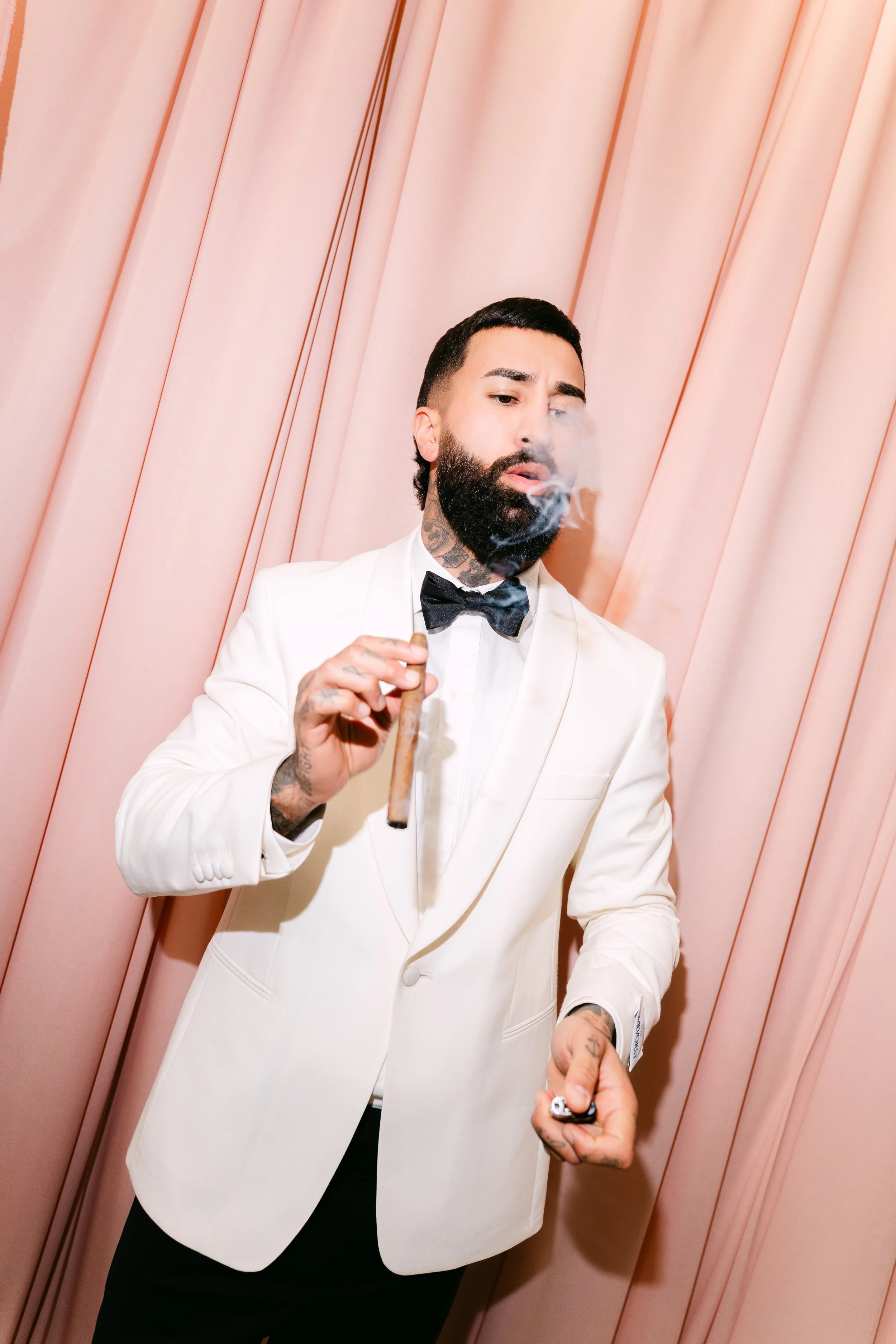A man with dark hair, a beard, and tattoos wearing a white tuxedo jacket, black bowtie, and black pants, smoking a cigar, standing in front of pink curtains.