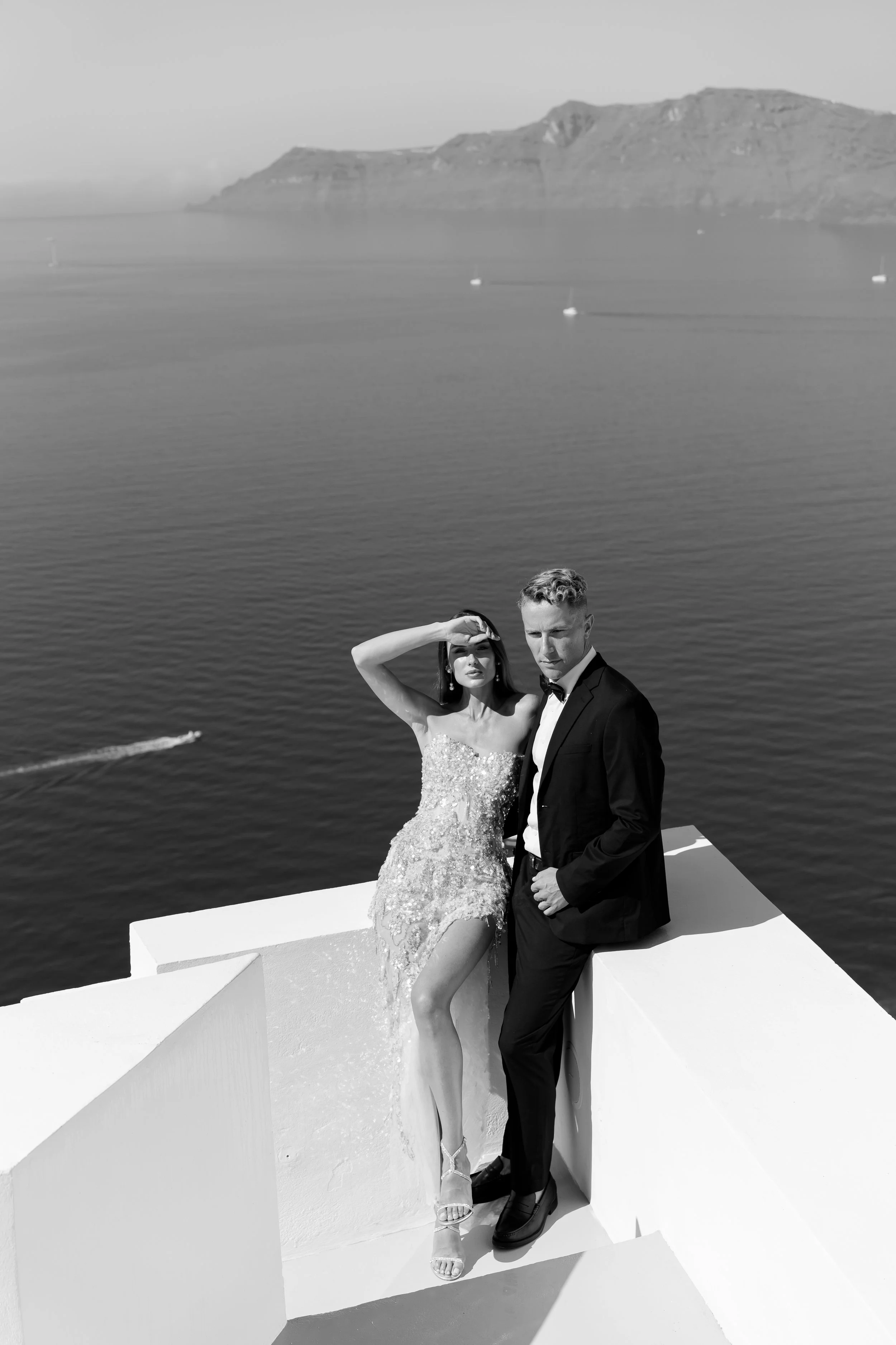 A black and white photo of a man and woman dressed formally, sitting on a white ledge by the water, with a mountainous island in the background.