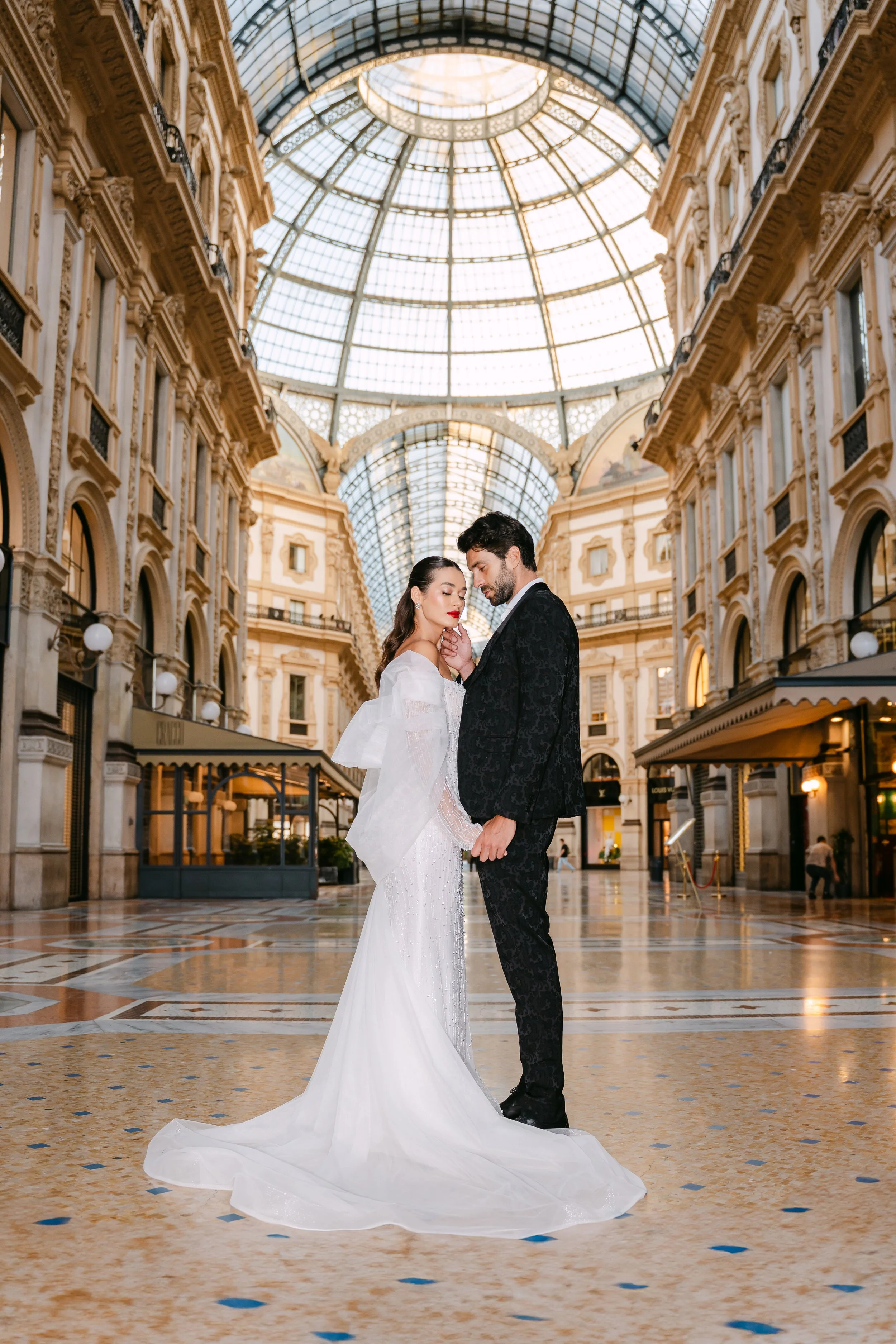 A bride and groom standing close together inside a grand, ornate shopping mall with a high glass-domed ceiling, elegant architecture, and warm lighting.