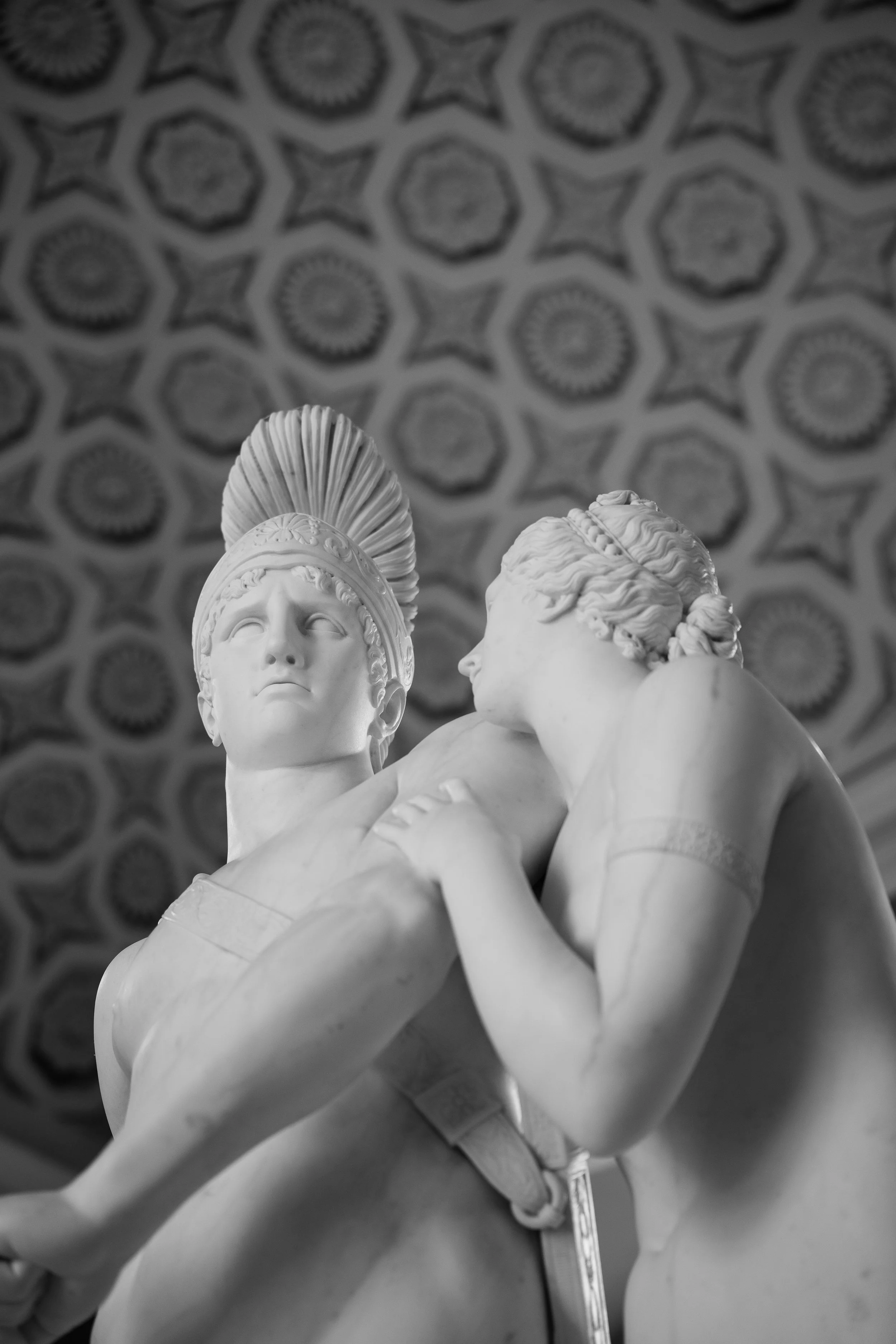 Close-up of a classical marble sculpture of two figures, a man with a helmet and a woman, against a patterned ceiling in the background.