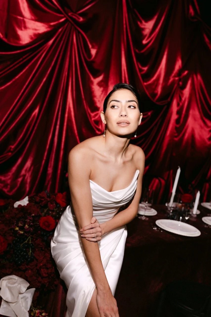 A woman in a white strapless dress sitting at a table with a dark red velvet curtain backdrop.