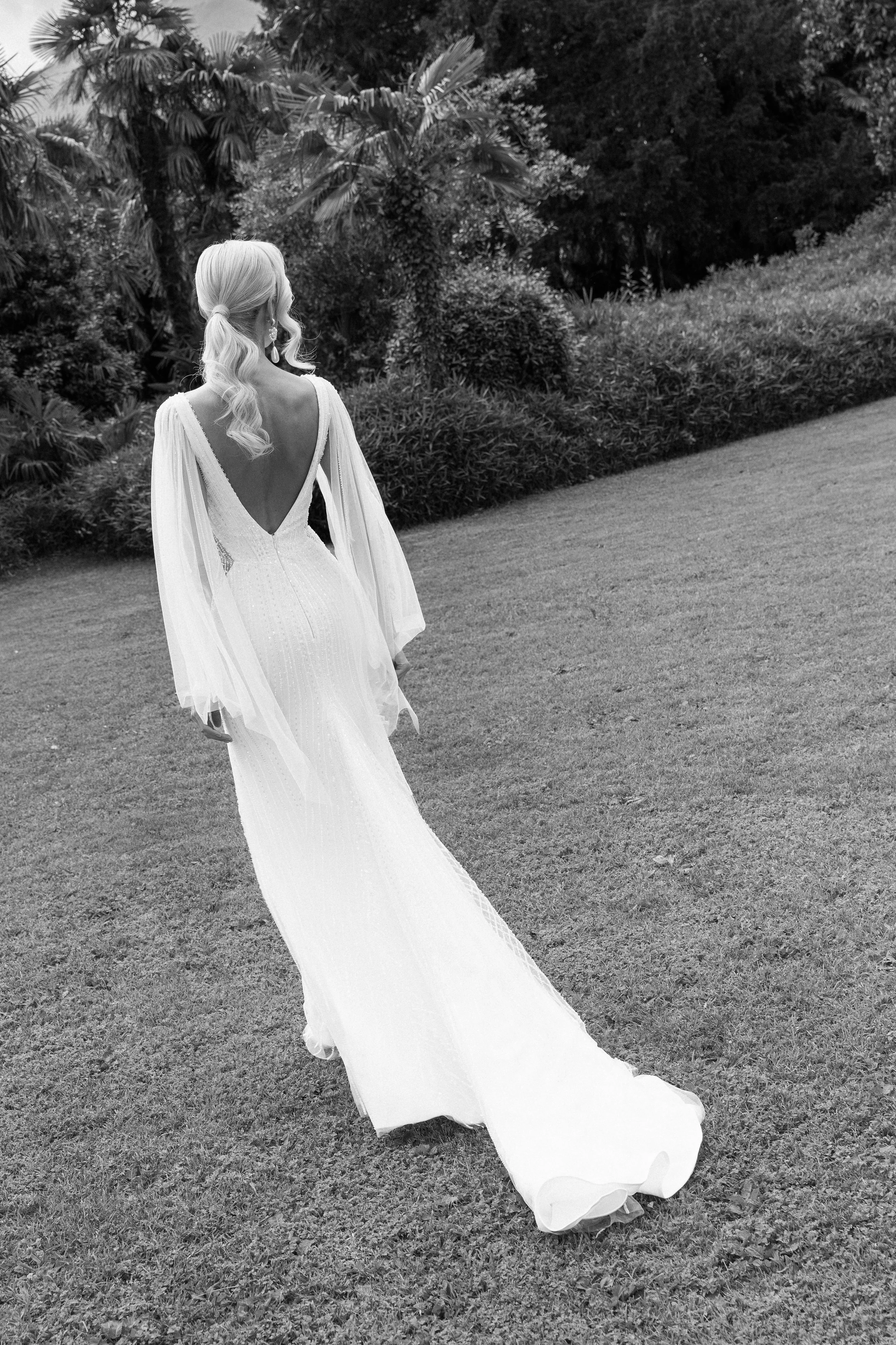 A woman in a long, flowing white dress with a deep V back, walking on grass in a garden with lush bushes and tall trees.