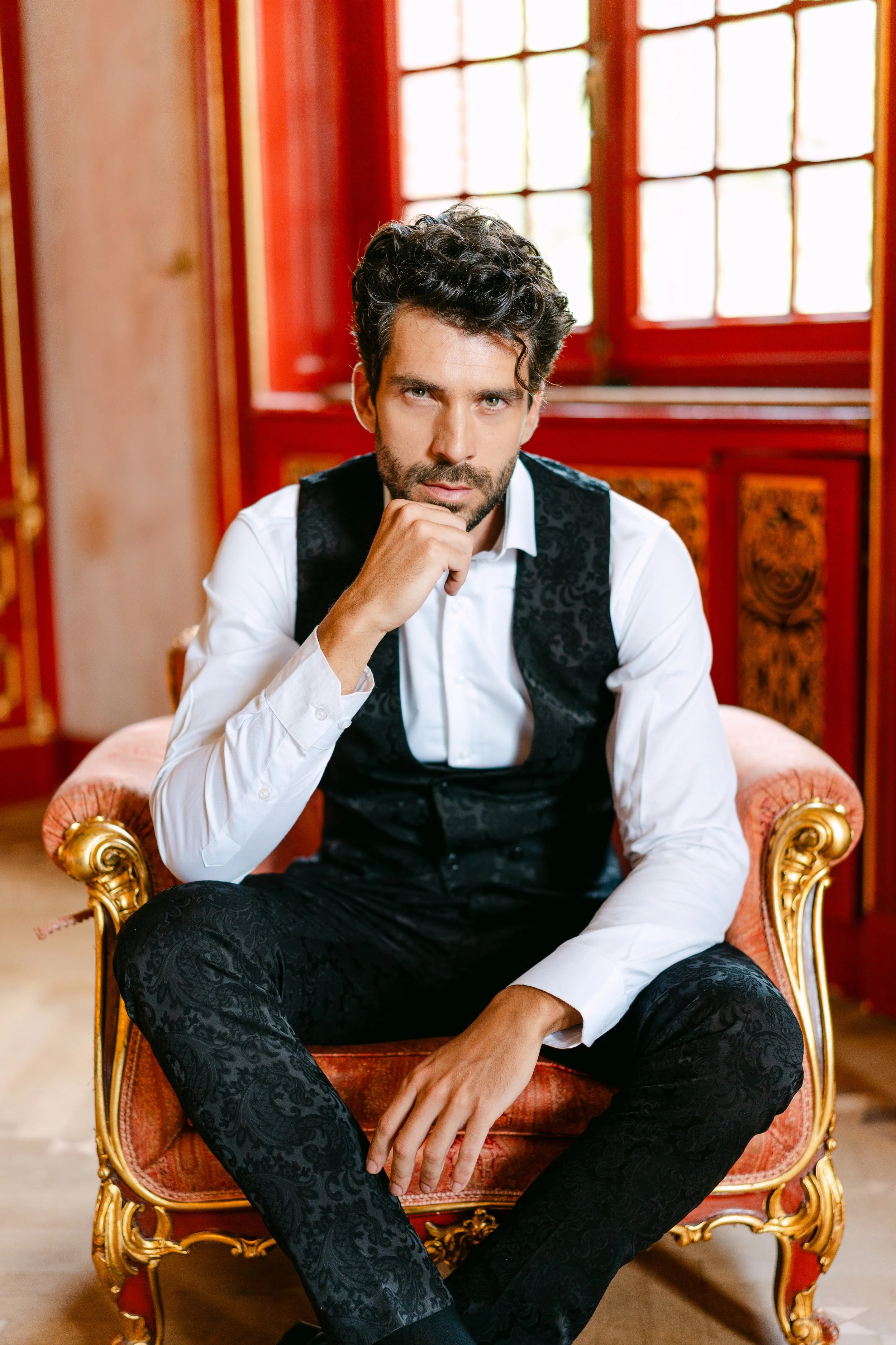 A man with dark curly hair and a beard, wearing a white dress shirt and a black patterned vest and pants, sitting on a vintage pink and gold upholstered armchair in a room with red wooden walls and large windows.
