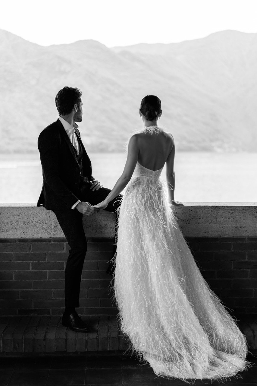 Black and white editorial wedding photography of a sophisticated couple overlooking Lake Como, Italy. The bride wears a luxury couture wedding dress with feathers and sequins, holding hands with her groom in a tailored suit against a scenic mountain 