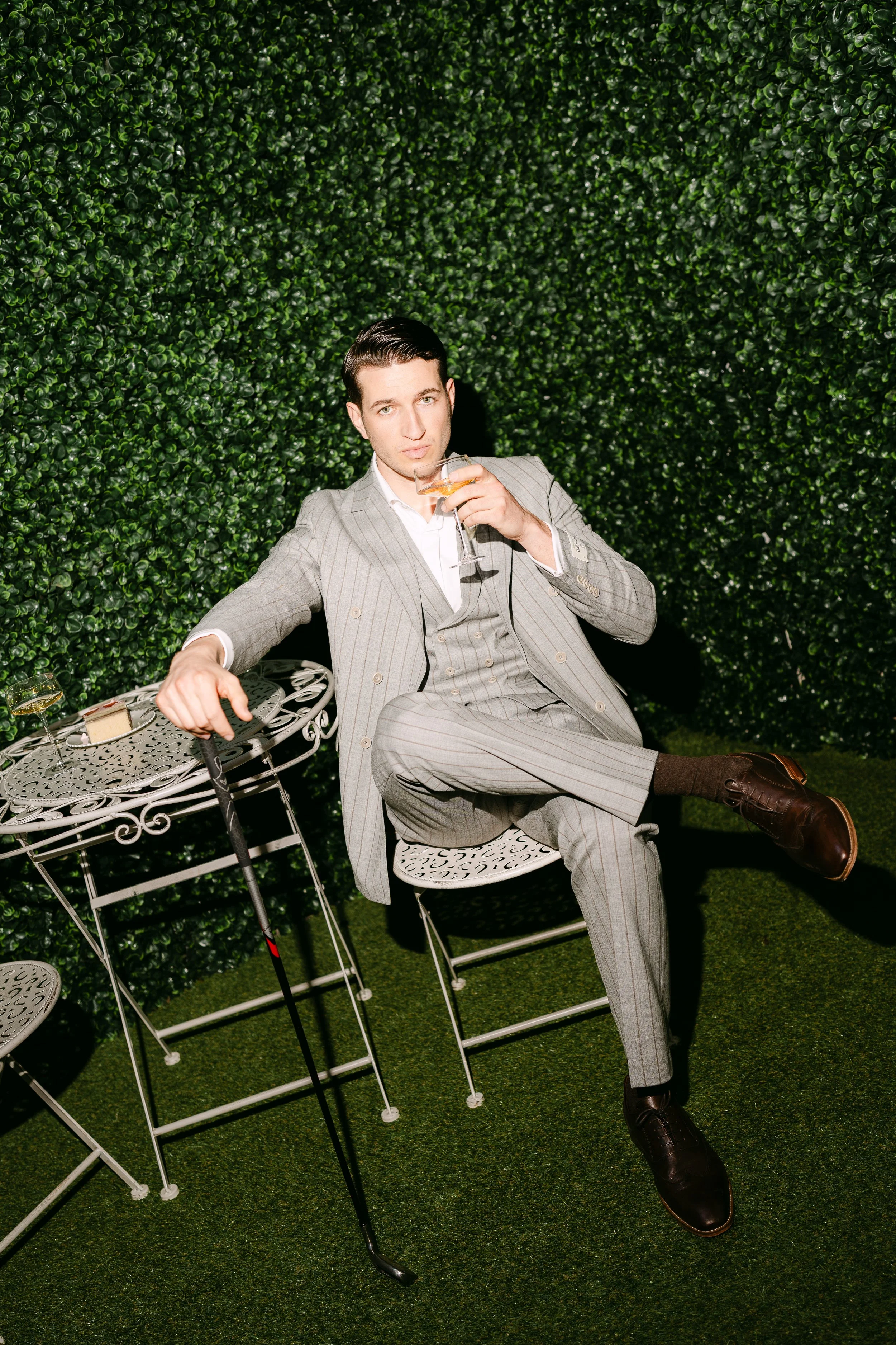 A man in a light gray pinstripe suit sitting outdoors on a white wrought iron chair, drinking from a wine glass, with a lush green hedge background, on a green grass surface.