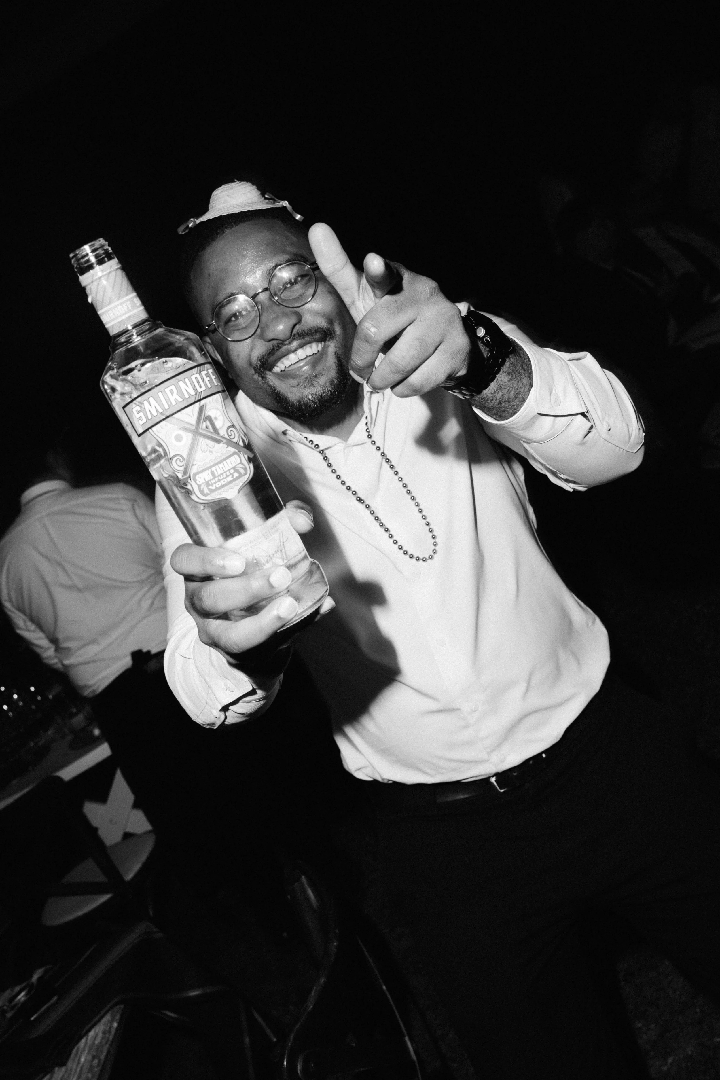 Man smiling and pointing at the camera, holding a bottle of Smirnoff vodka at a party or social gathering, in black and white.
