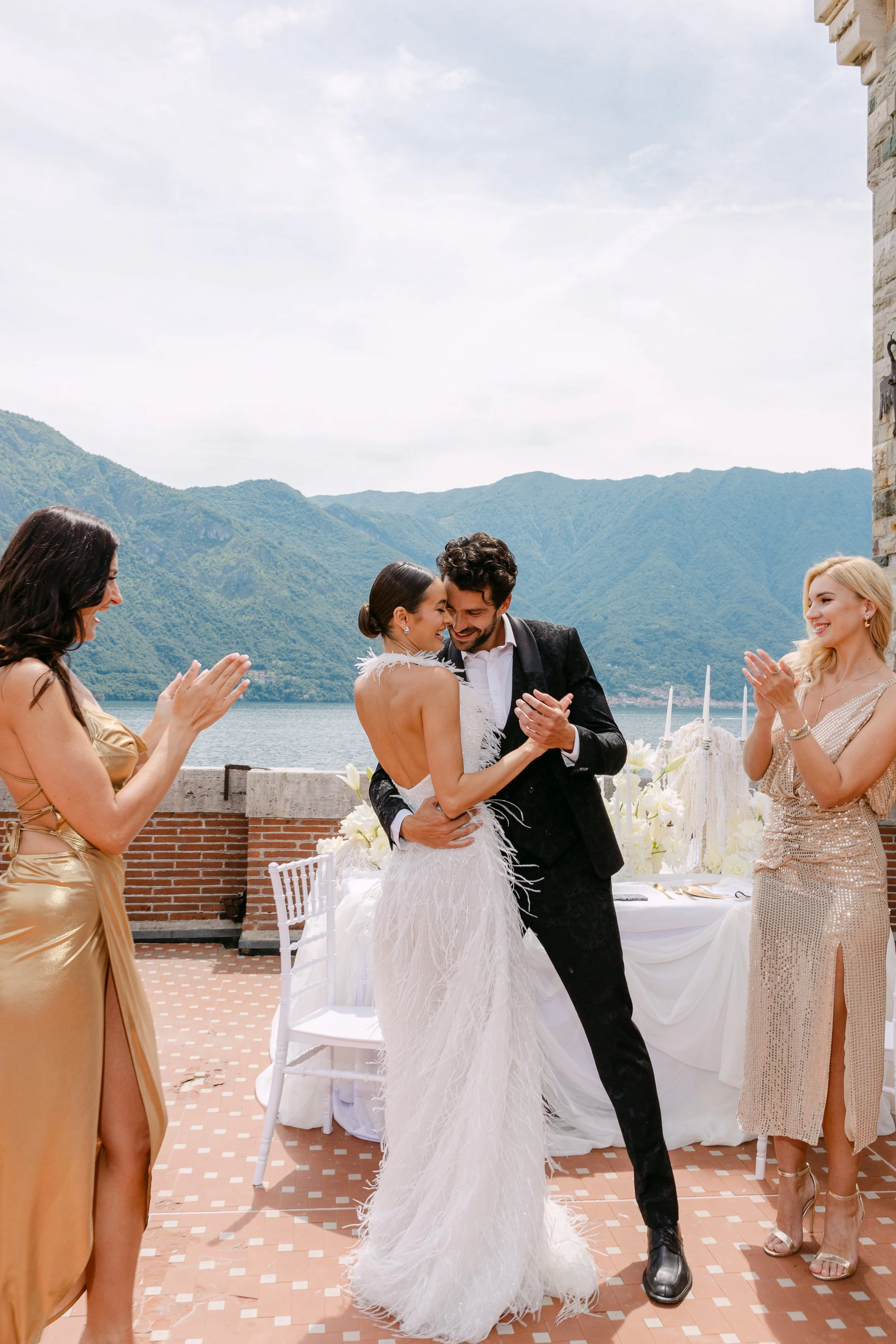 A couple's wedding dance outdoors by a lake with mountain views, surrounded by four women clapping and celebrating.