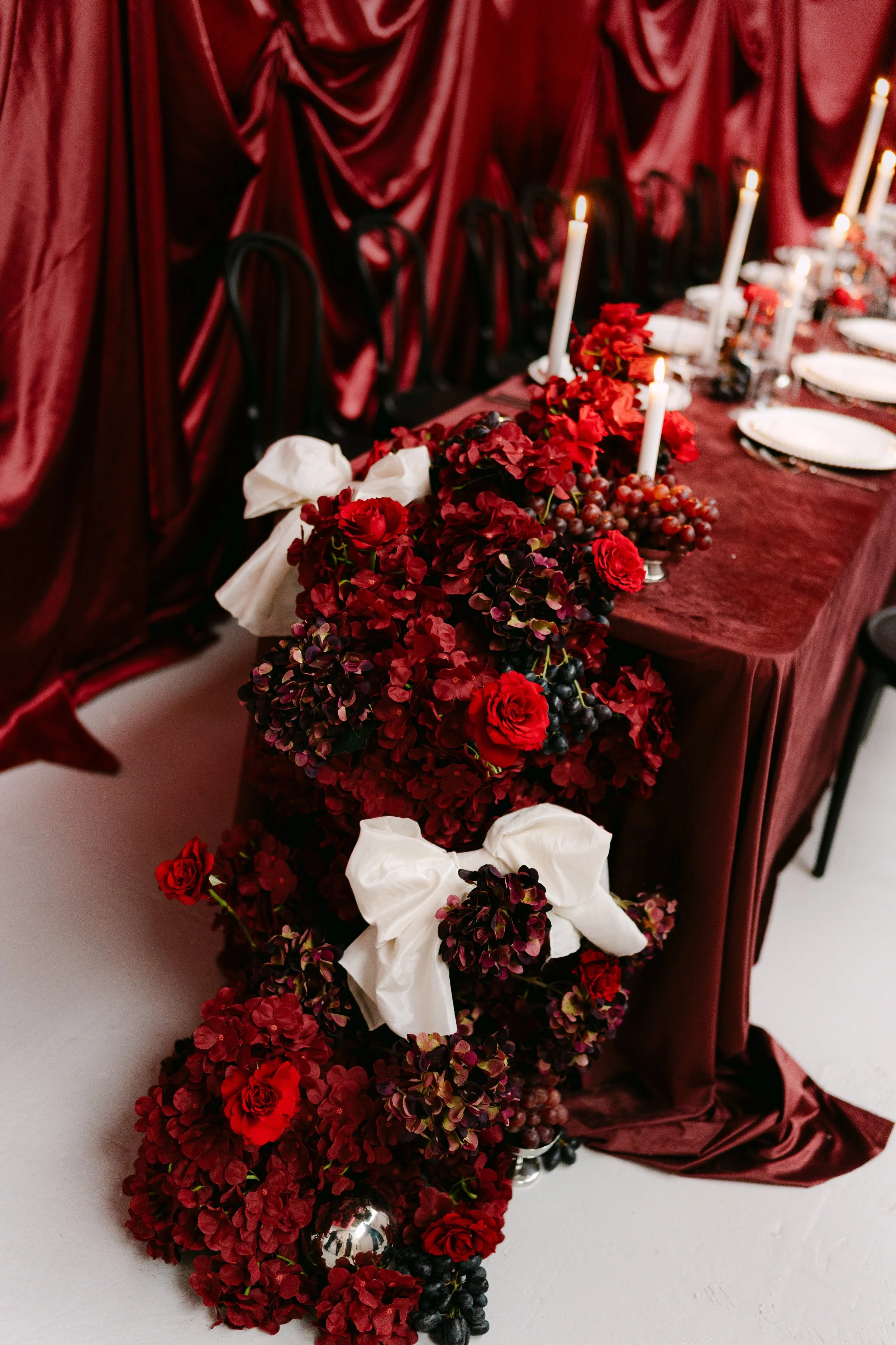 A table decorated for a formal event with a red floral centerpiece, white candles, and elegant tableware, with a matching red draped tablecloth and backdrop.