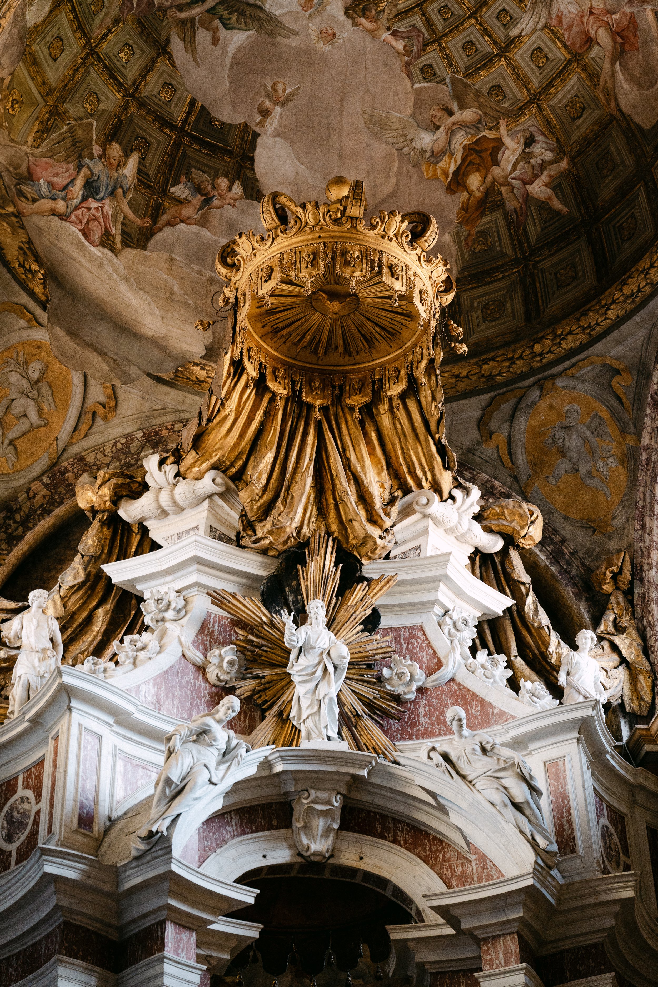 Baroque altar with white statues of saints and angels, gold drapery decoration, and a painted ceiling with cherubs and clouds.