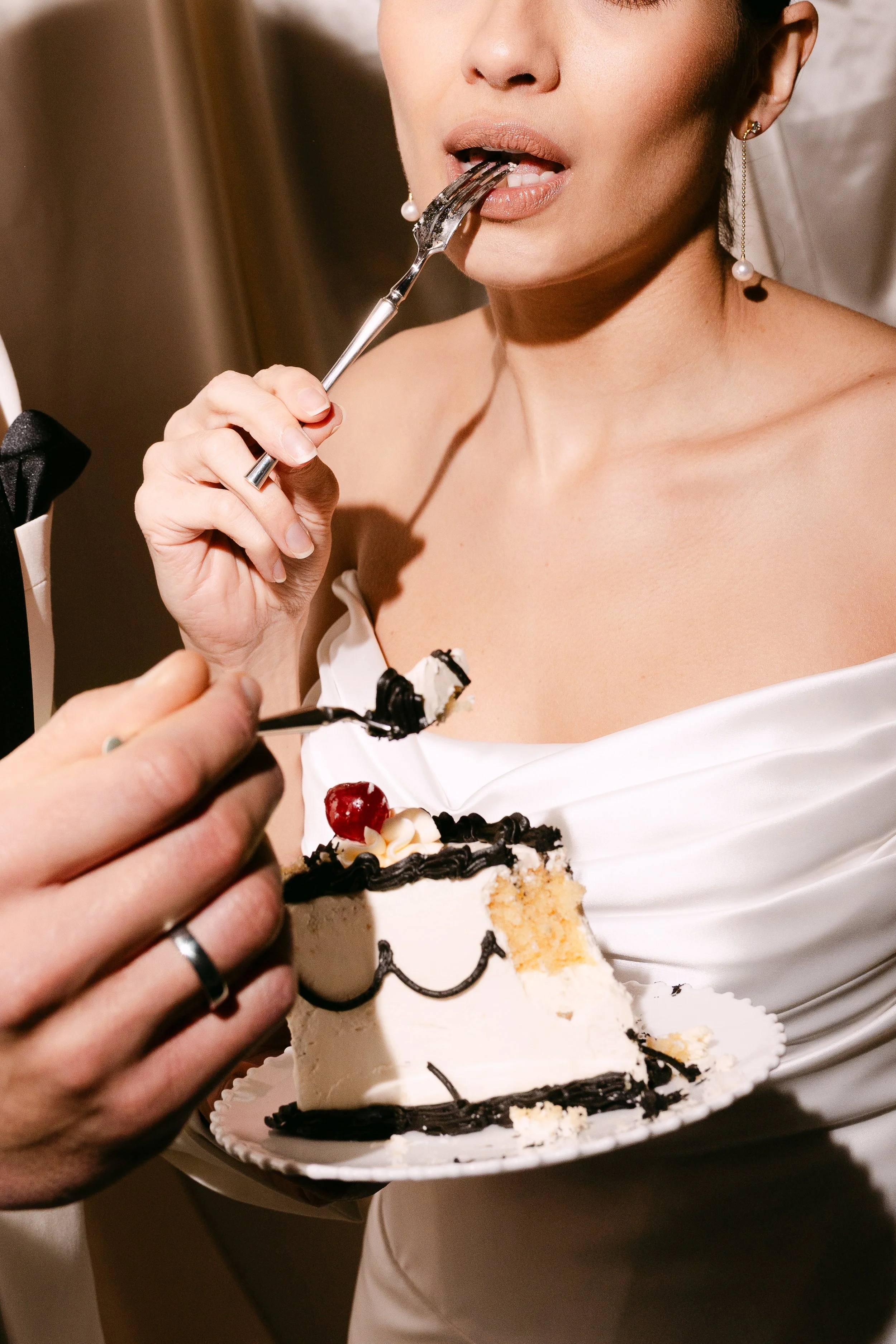 Close-up of a bride in a white dress and pearl earrings, about to eat a slice of wedding cake with a fork, as someone helps serve it.