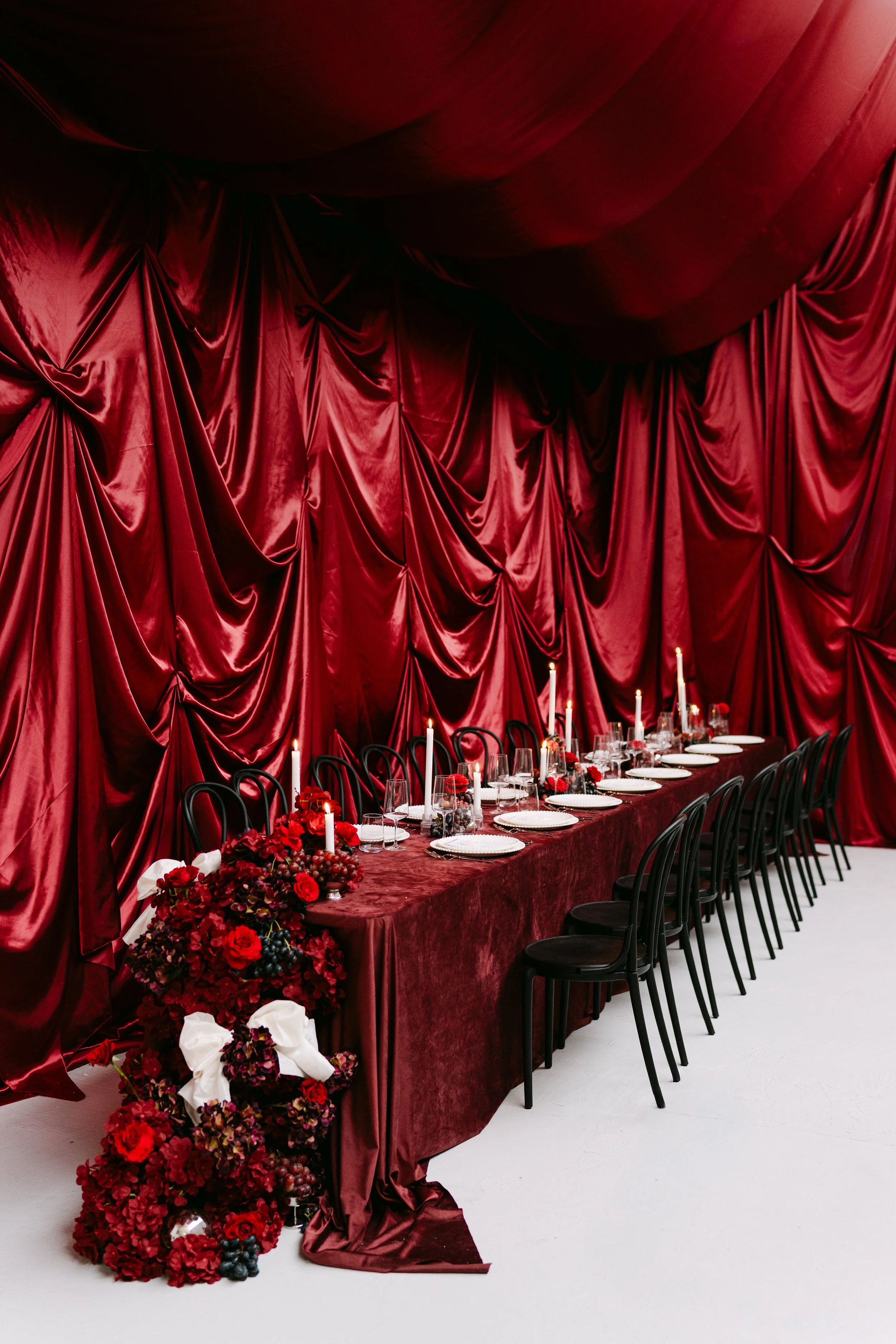 A long banquet table set for a formal event against a red velvet draped backdrop, with black chairs, white plates, glassware, tall candles, and red floral centerpieces.