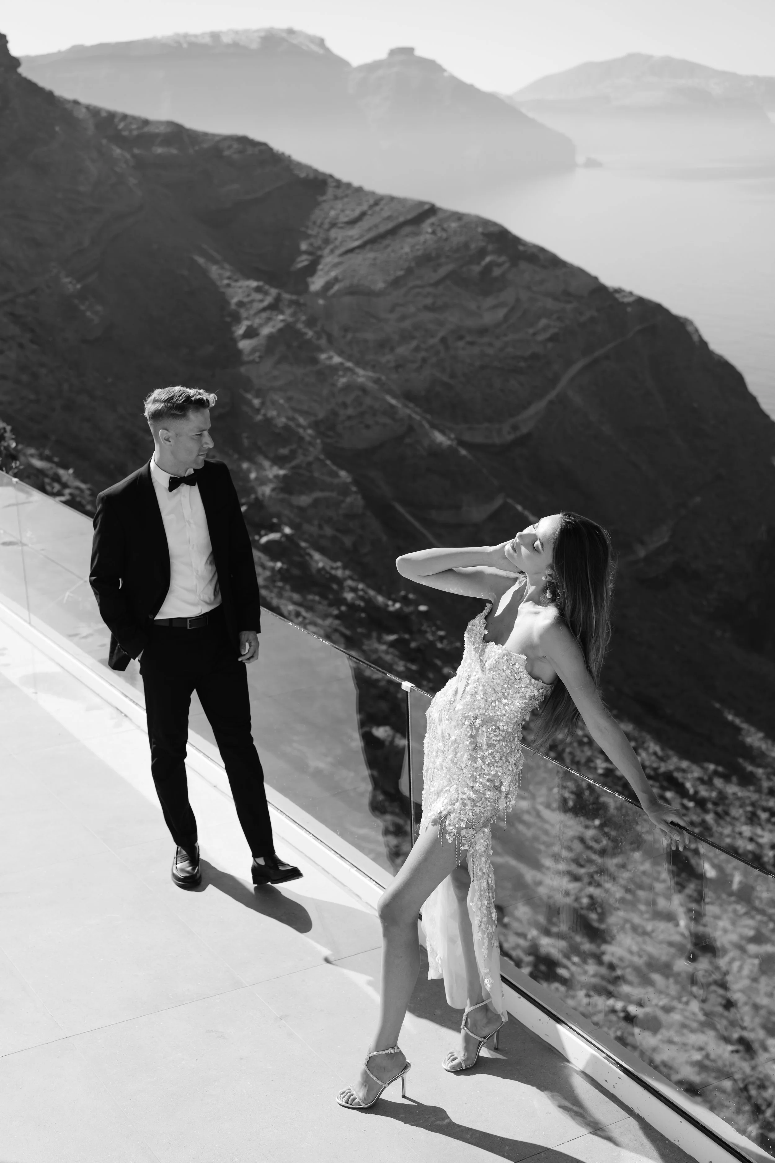 A black and white photo of a man in a tuxedo and woman in an elegant dress on a balcony overlooking mountains and water.