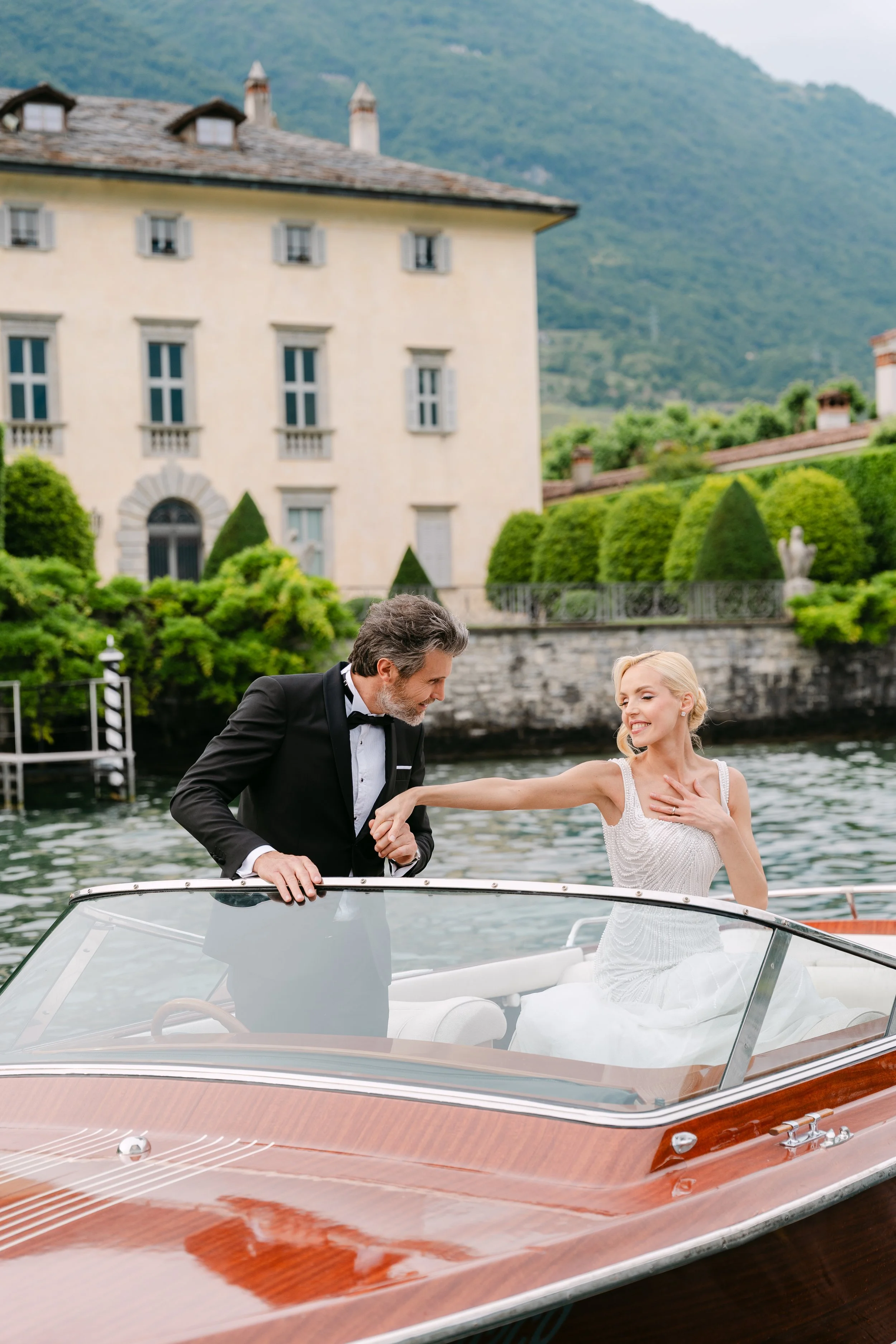 A groom in a black tuxedo helps a bride in a white wedding dress onto a boat on a lake, with a historic house and lush green hills in the background.