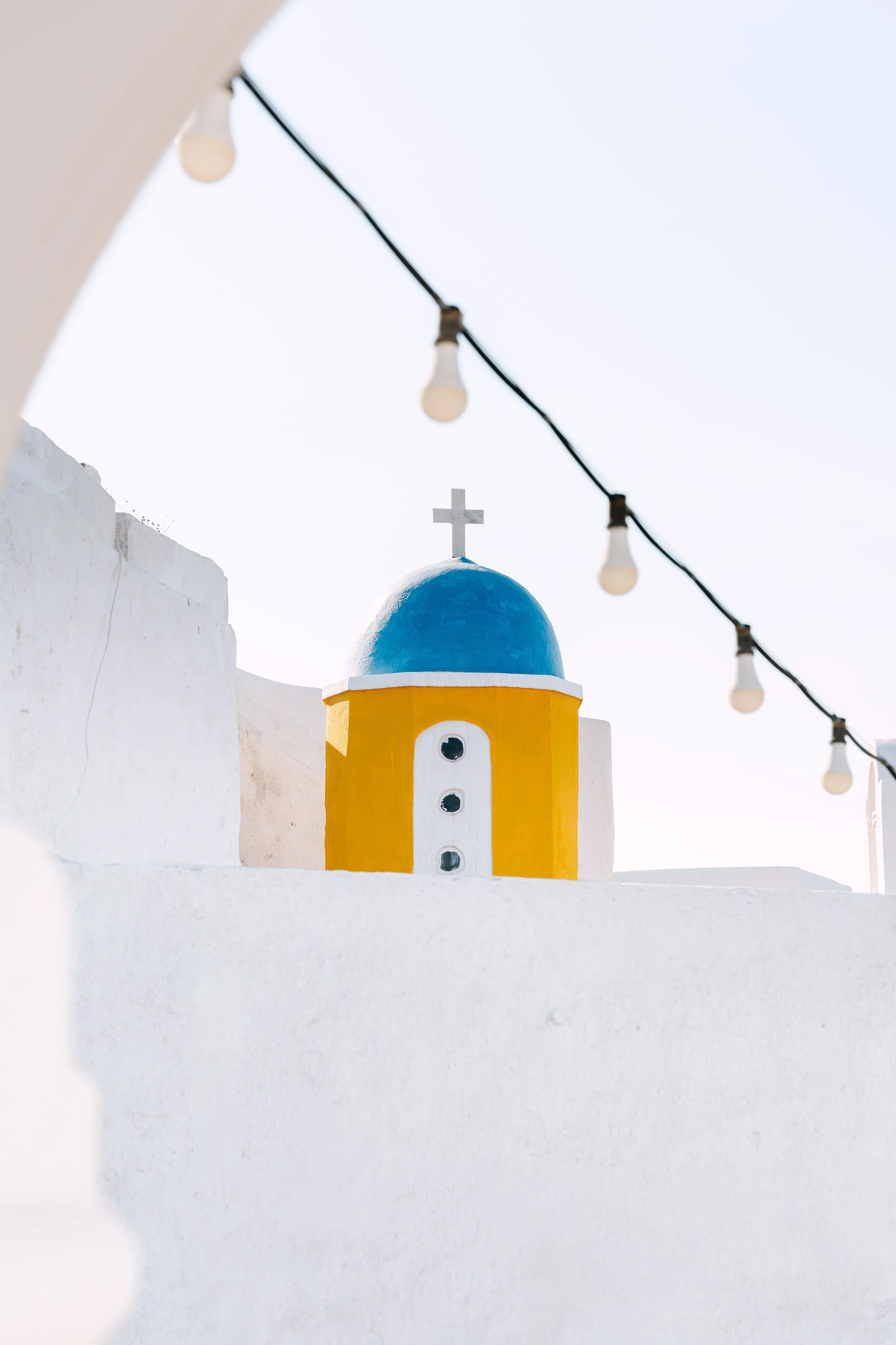A white building with a blue dome and a cross on top, decorated with a yellow and white facade, in a sunny setting with string lights overhead.