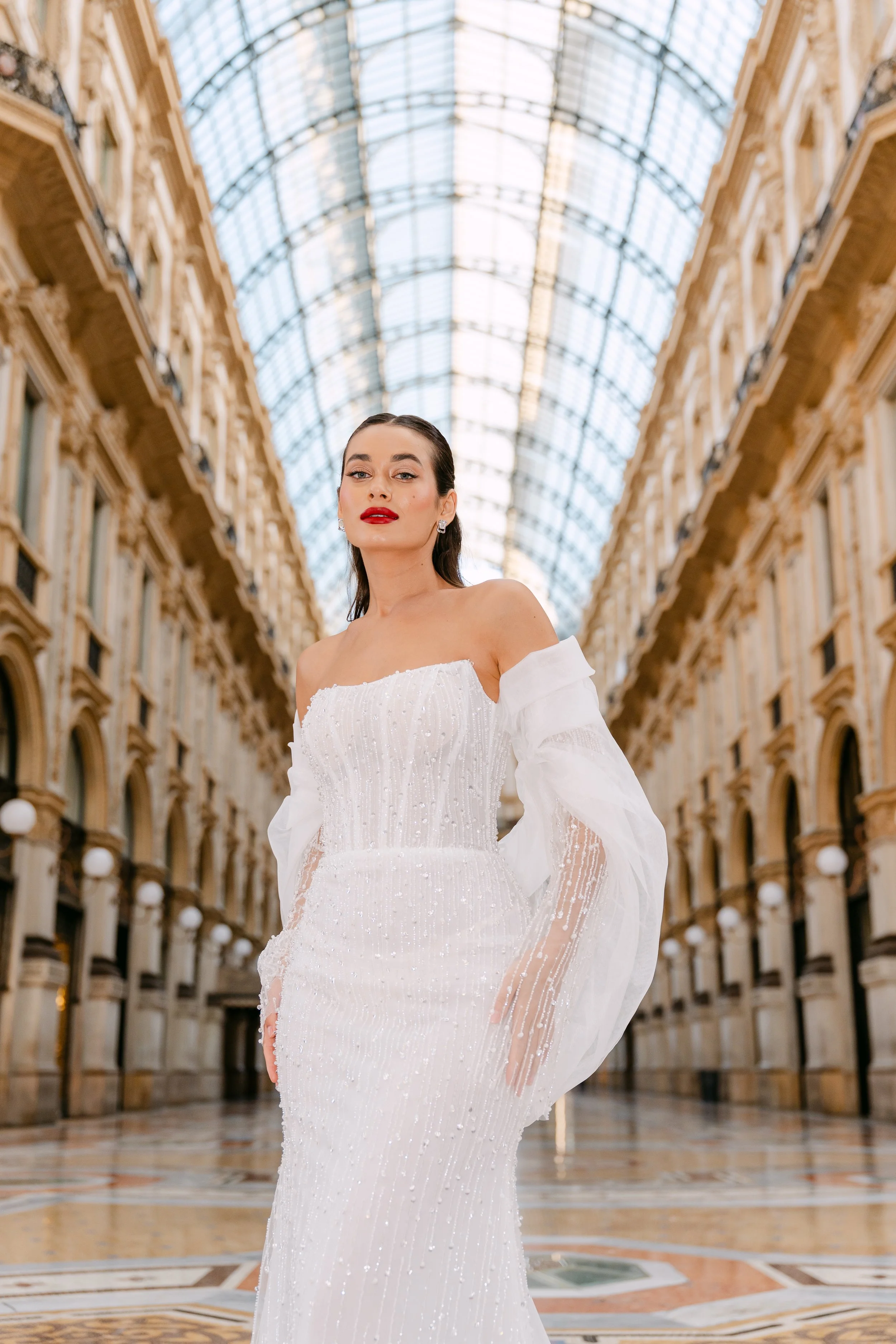 A woman in a white dress with sheer, puffy sleeves standing in an ornate, glass-ceilinged shopping arcade.