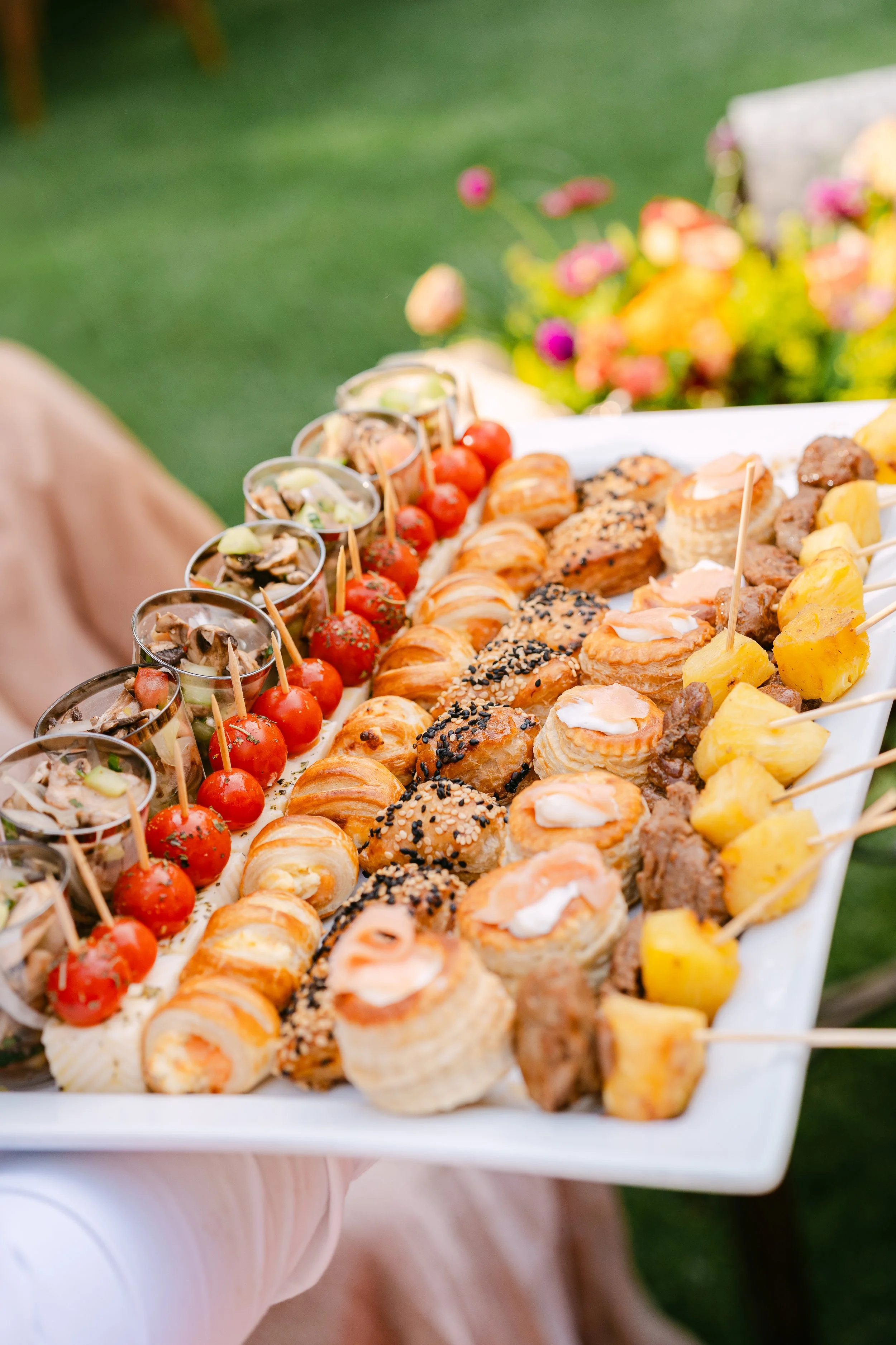 Assorted appetizers including cherry tomatoes, scallop cups, mini croissants, sesame seed-topped pastries, shrimp canapés, and pineapple chunks on a white serving tray held outside with green grass and colorful flowers in the background.