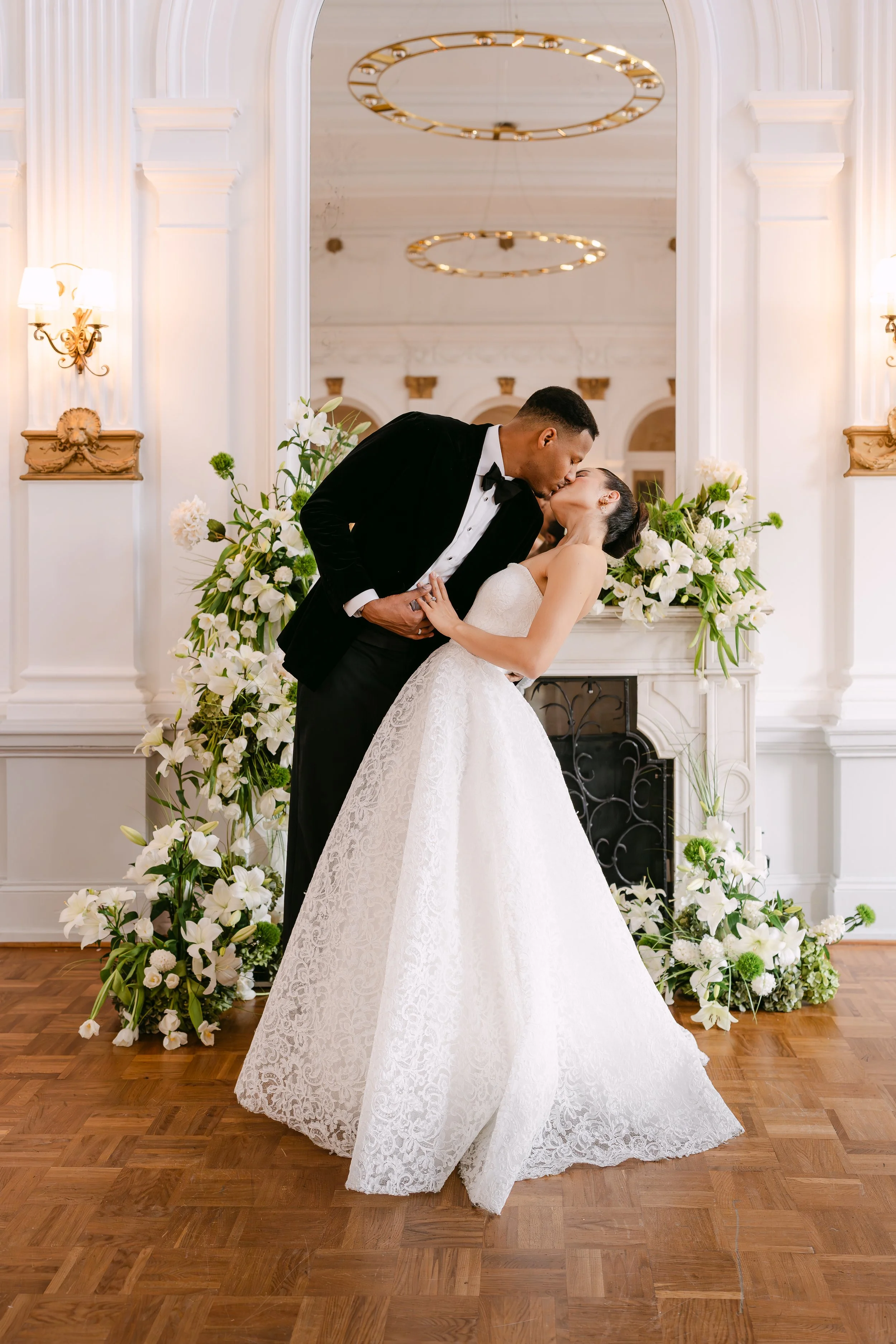 A newlywed couple sharing a kiss during their wedding ceremony in an elegant, decorated room with floral arrangements and wooden flooring.