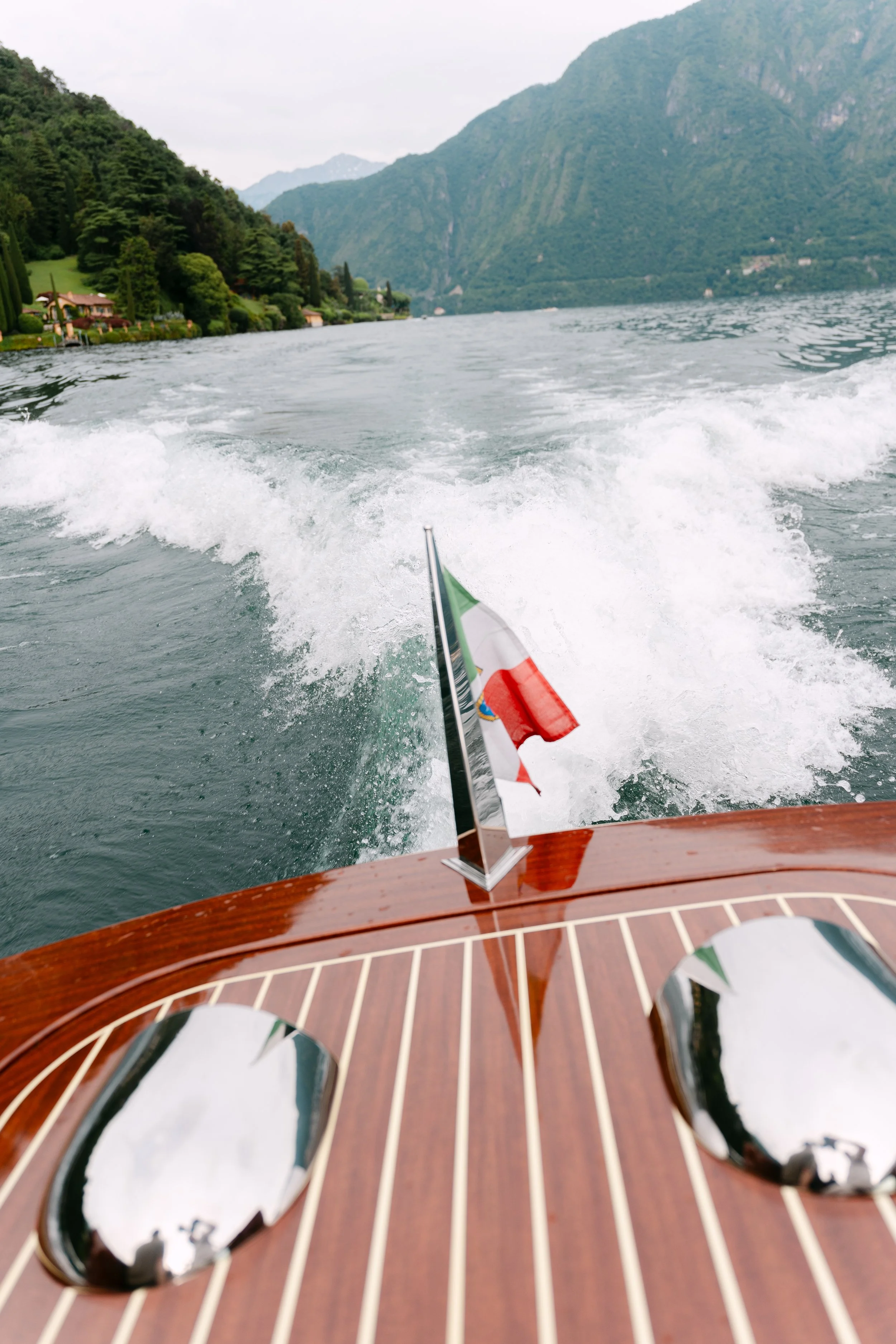View from a boat on a lake with mountains in the background, showing water spray and small Italian flag on the boat's wooden surface.