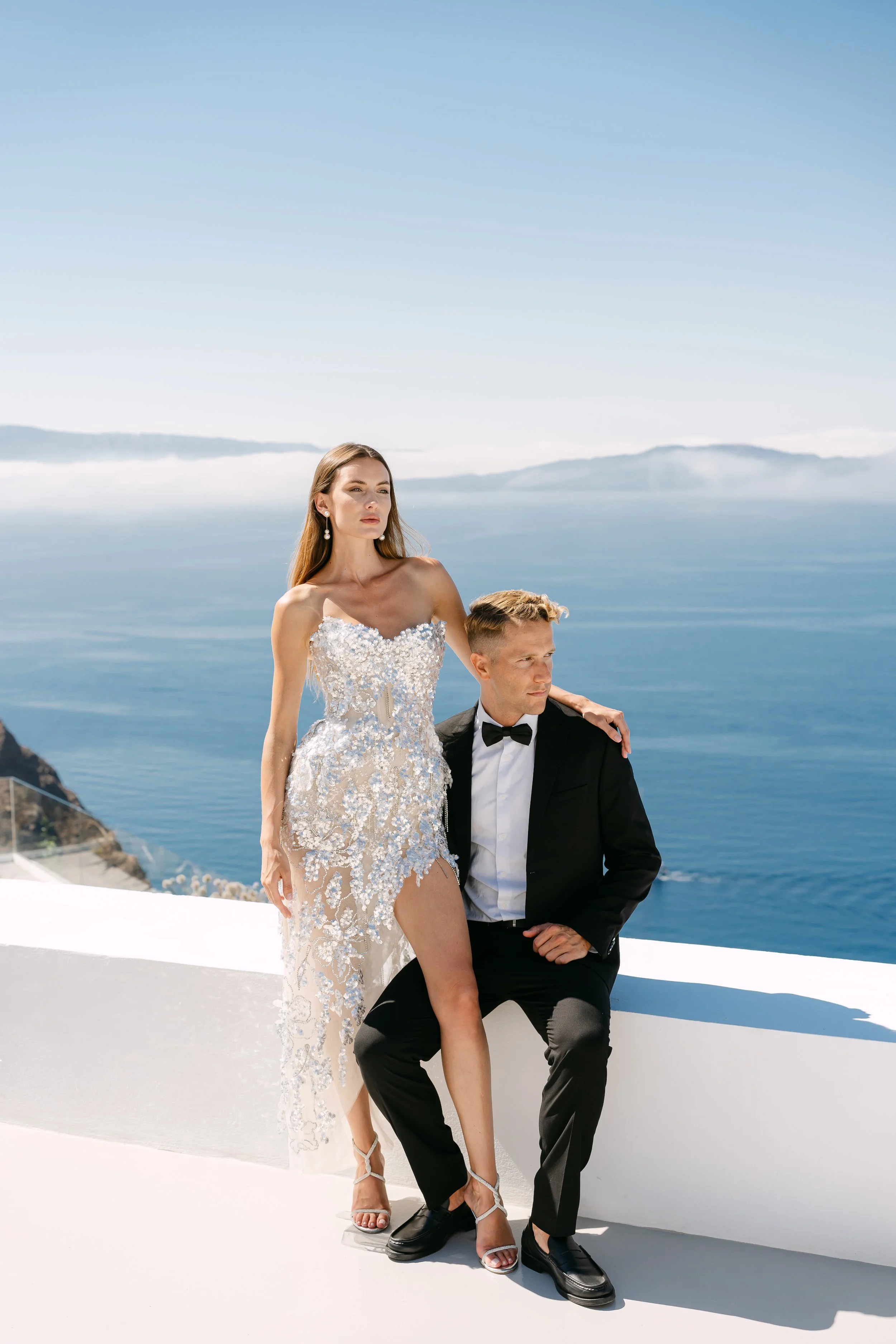 A woman in a detailed, sparkling white wedding dress and heels stands with her hand on a seated man in a tuxedo, against a background of the sea and sky.