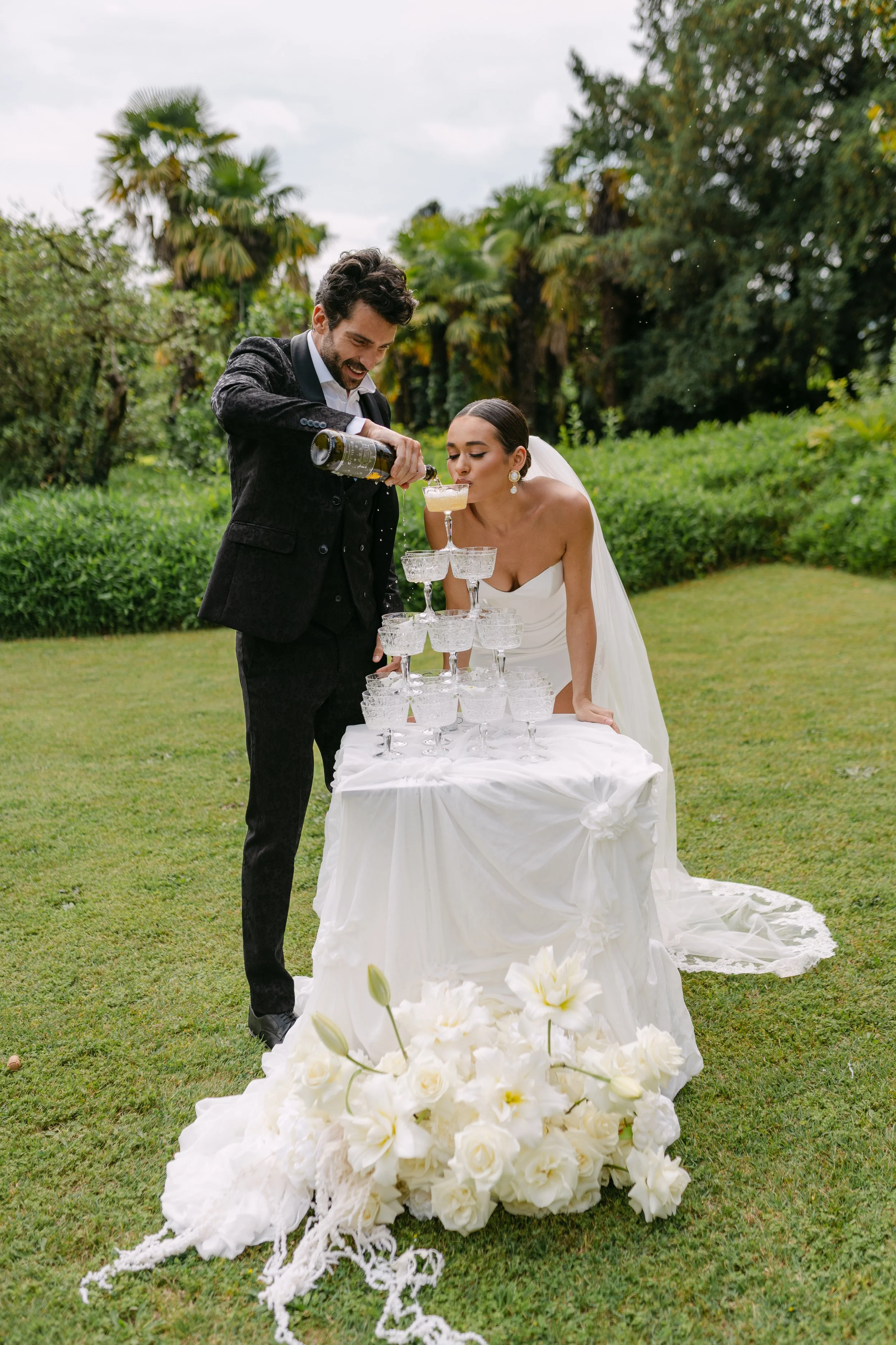 Bride and groom celebrate and pour champagne into a tower of glasses at an outdoor wedding reception surrounded by greenery and flowers.