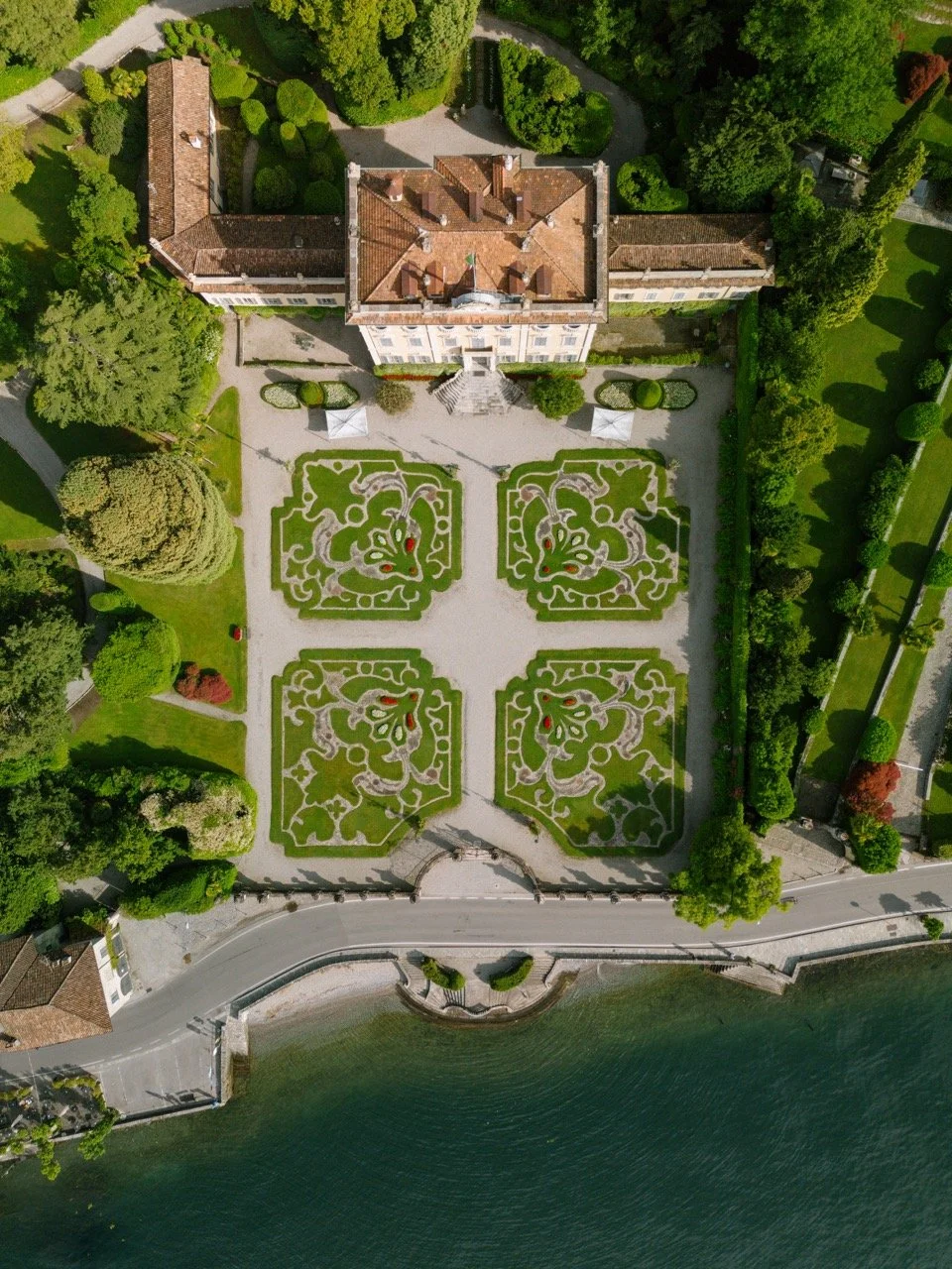 An aerial view of a grand estate with formal garden lawns, a large building with red-tiled roof, and a waterfront area with a curved walkway.