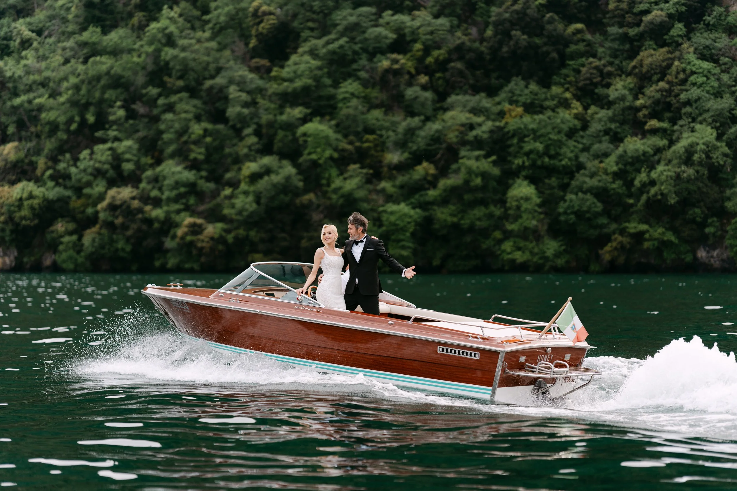 A couple dressed in wedding attire riding a wooden speedboat on a lake with a forested background.