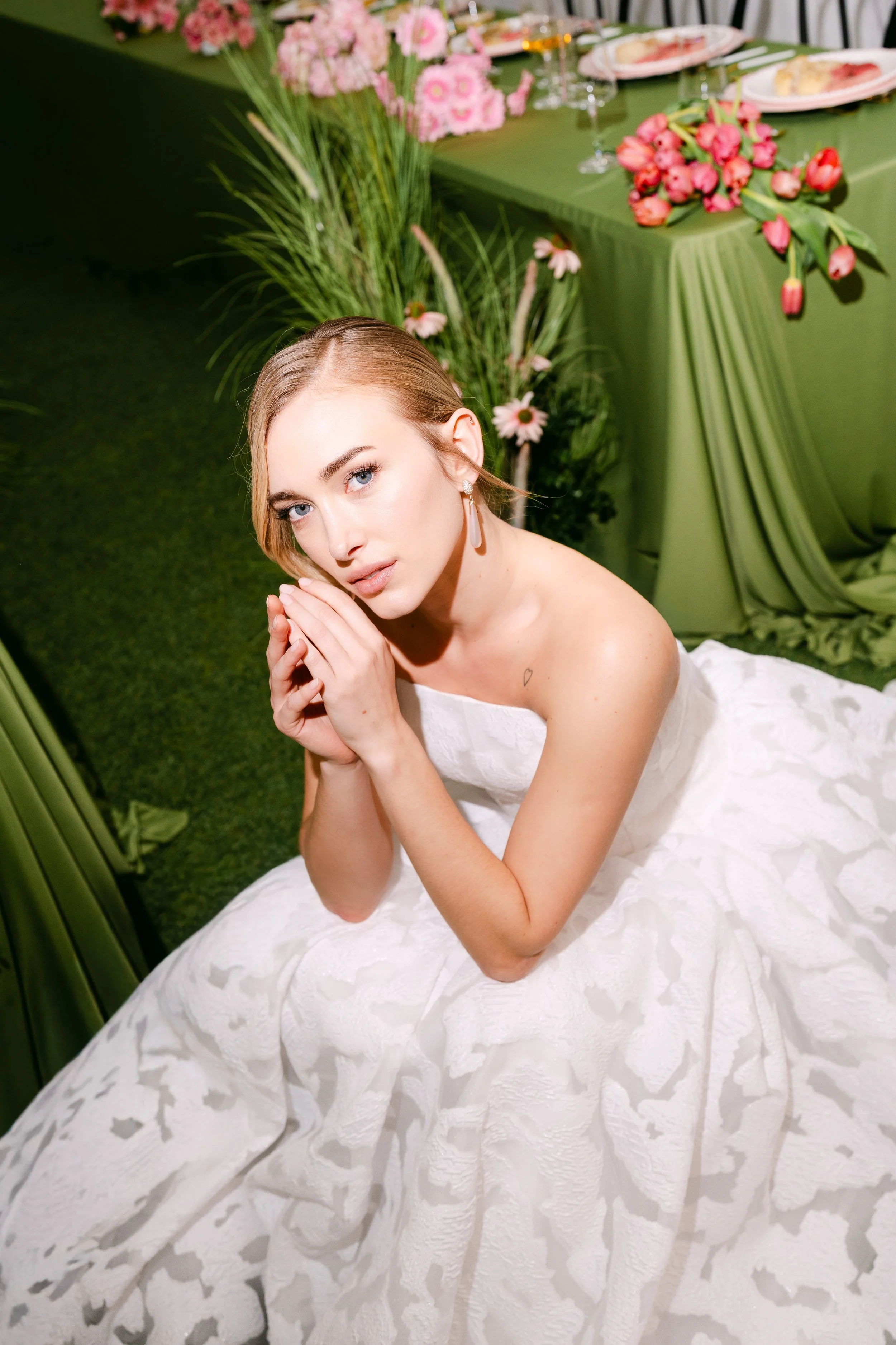 A woman with blonde hair and blue eyes, dressed in a white textured dress, is sitting on a dress at a decorated event table with pink flowers, green tablecloth, and dinnerware in the background.