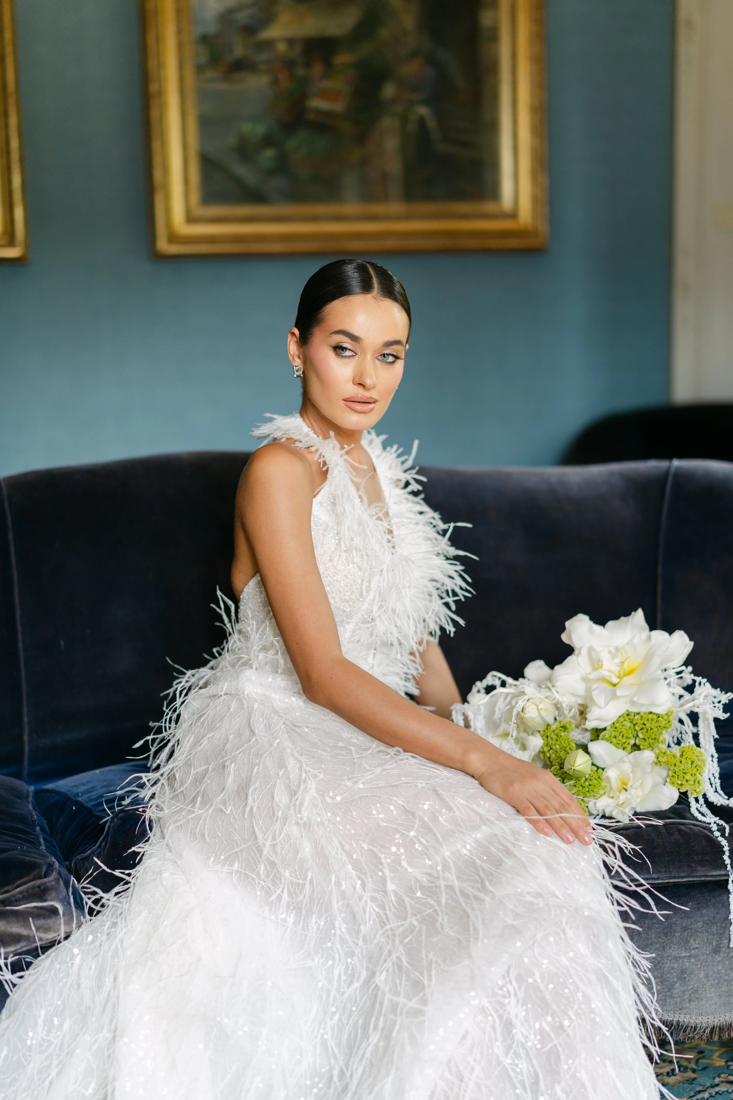 A woman in a white, feathered wedding dress sitting on a dark velvet sofa, holding a bouquet of white and green flowers, with framed paintings on a blue wall behind her.